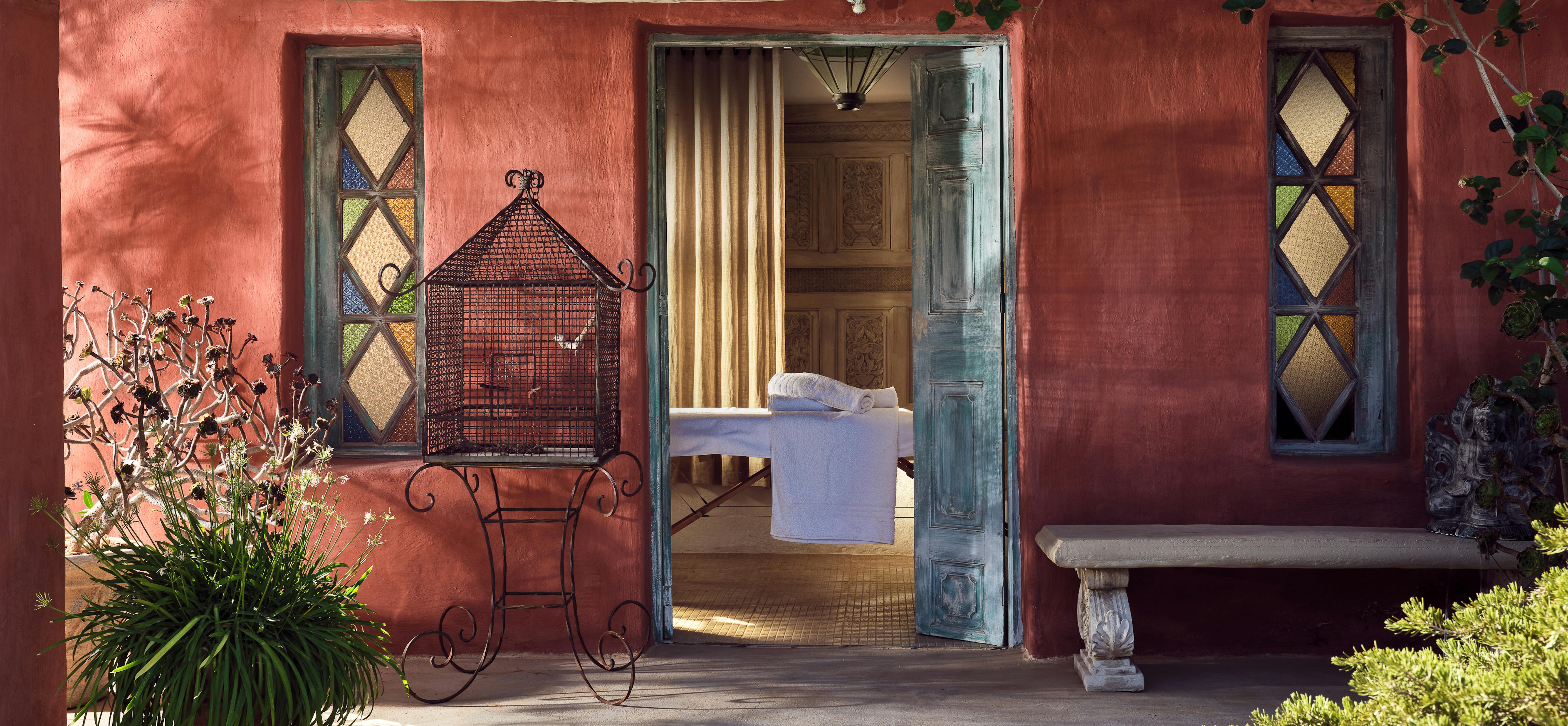 Covered terrace entrance with coral-red walls, colourful stained glass windows, ornate metal birdcage chair, weathered blue door opening to bedroom.