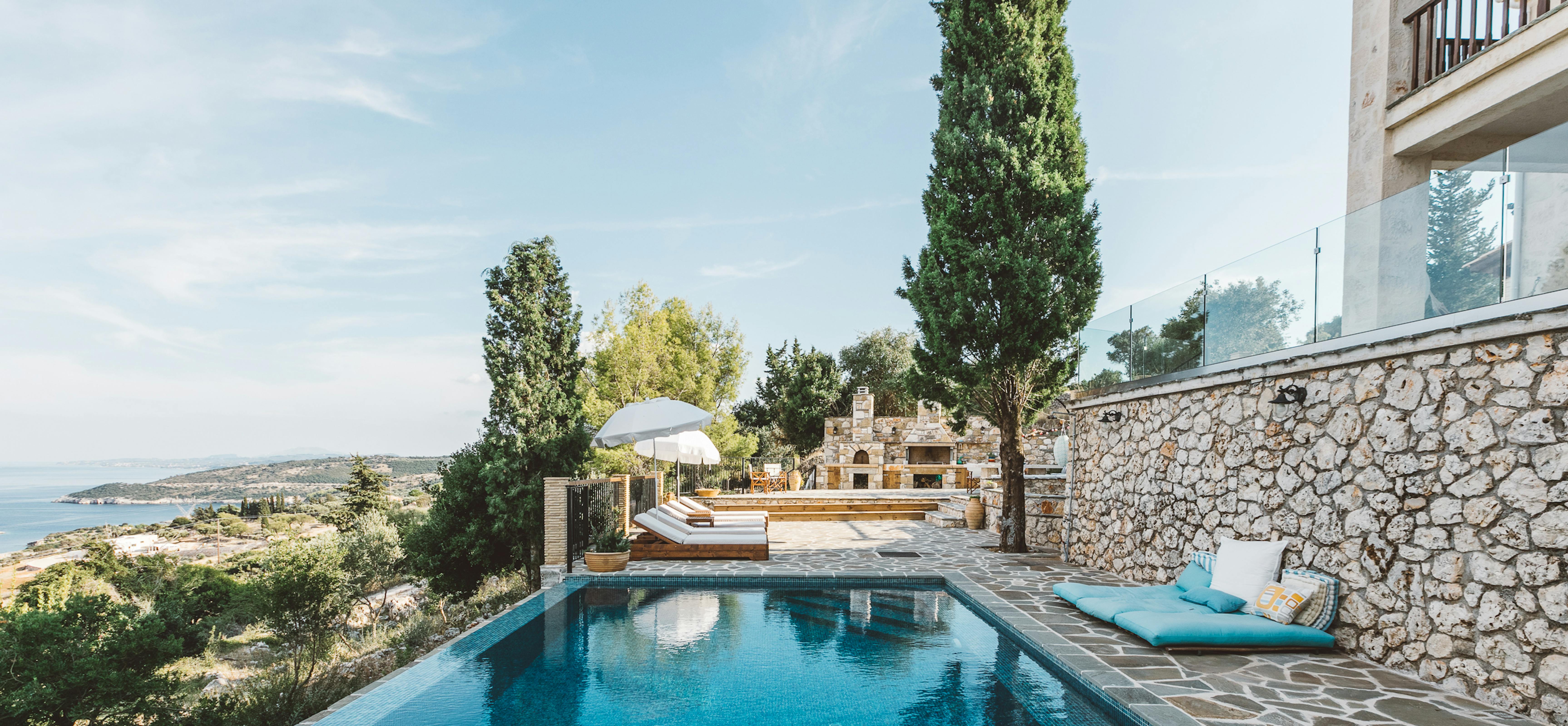 Hillside infinity pool with mosaic tile finish overlooking coastal landscape, stone terrace with turquoise cushioned seating, white umbrellas, cypress trees, and Mediterranean villa.