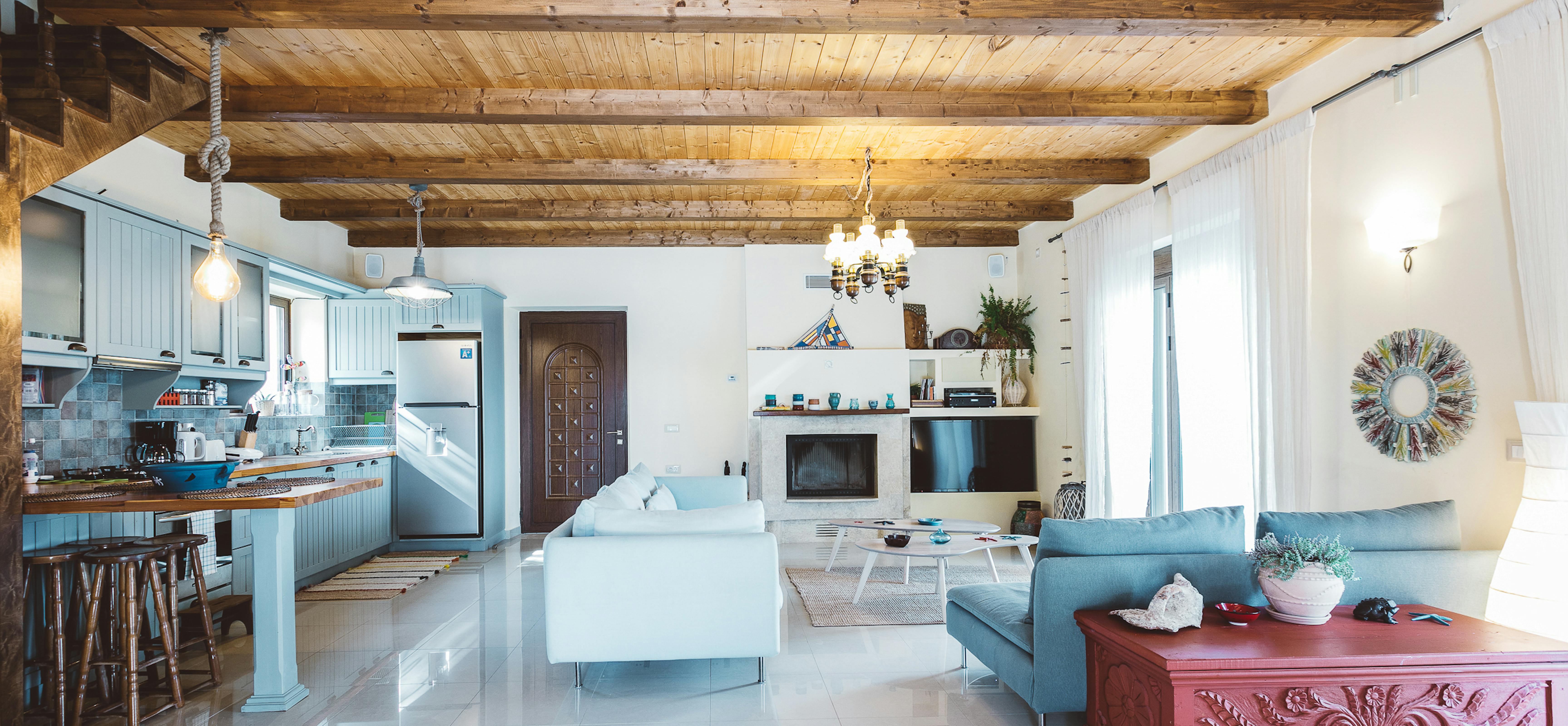 Open-concept living space with exposed wood beam ceiling, white sofas with turquoise accents, pale blue kitchen, glossy white floors, and ornate red carved chest.