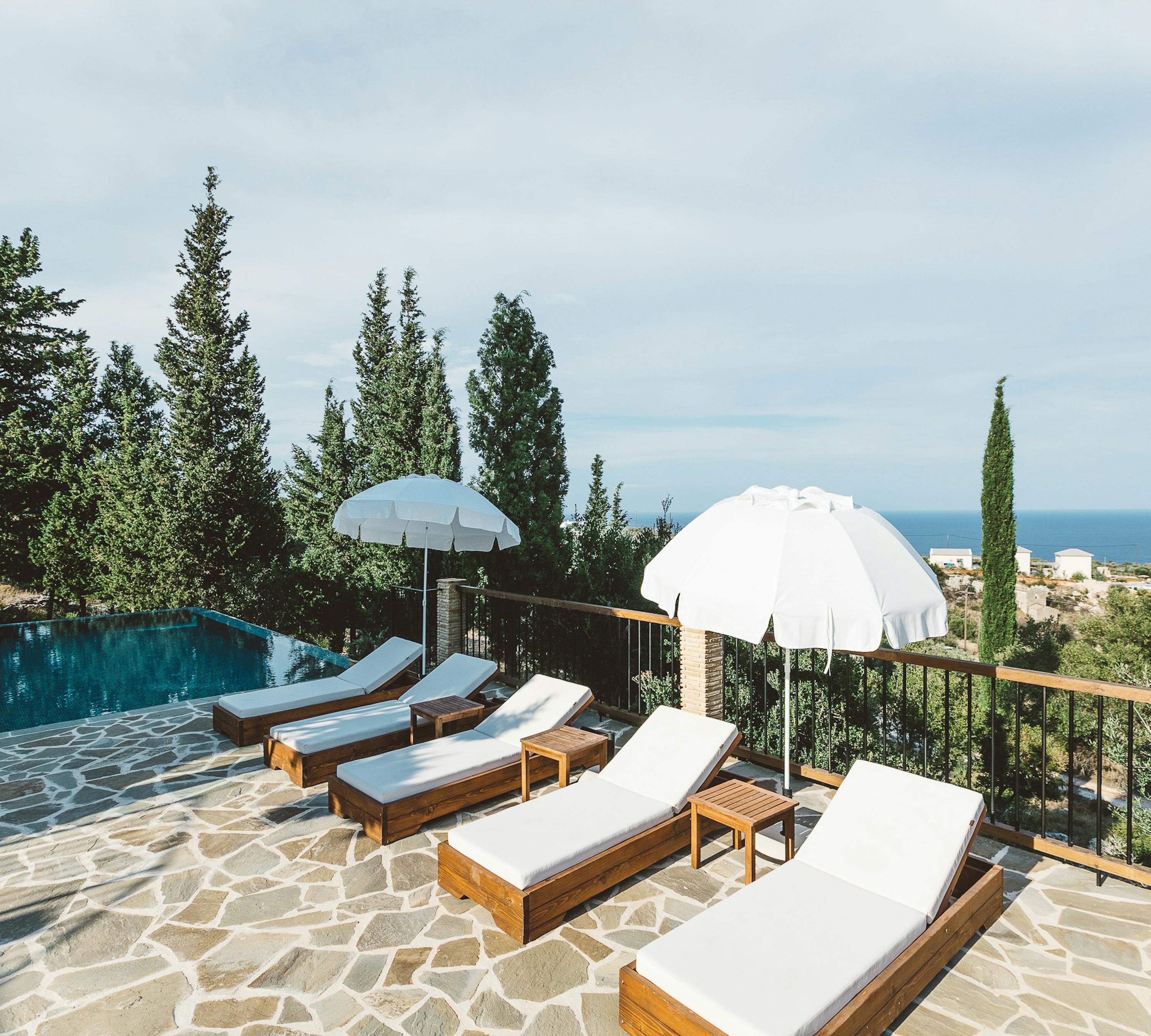 Pool terrace with wooden loungers, white umbrella, irregular stone paving, glass safety railing, cypress trees, and distant sea and town views.