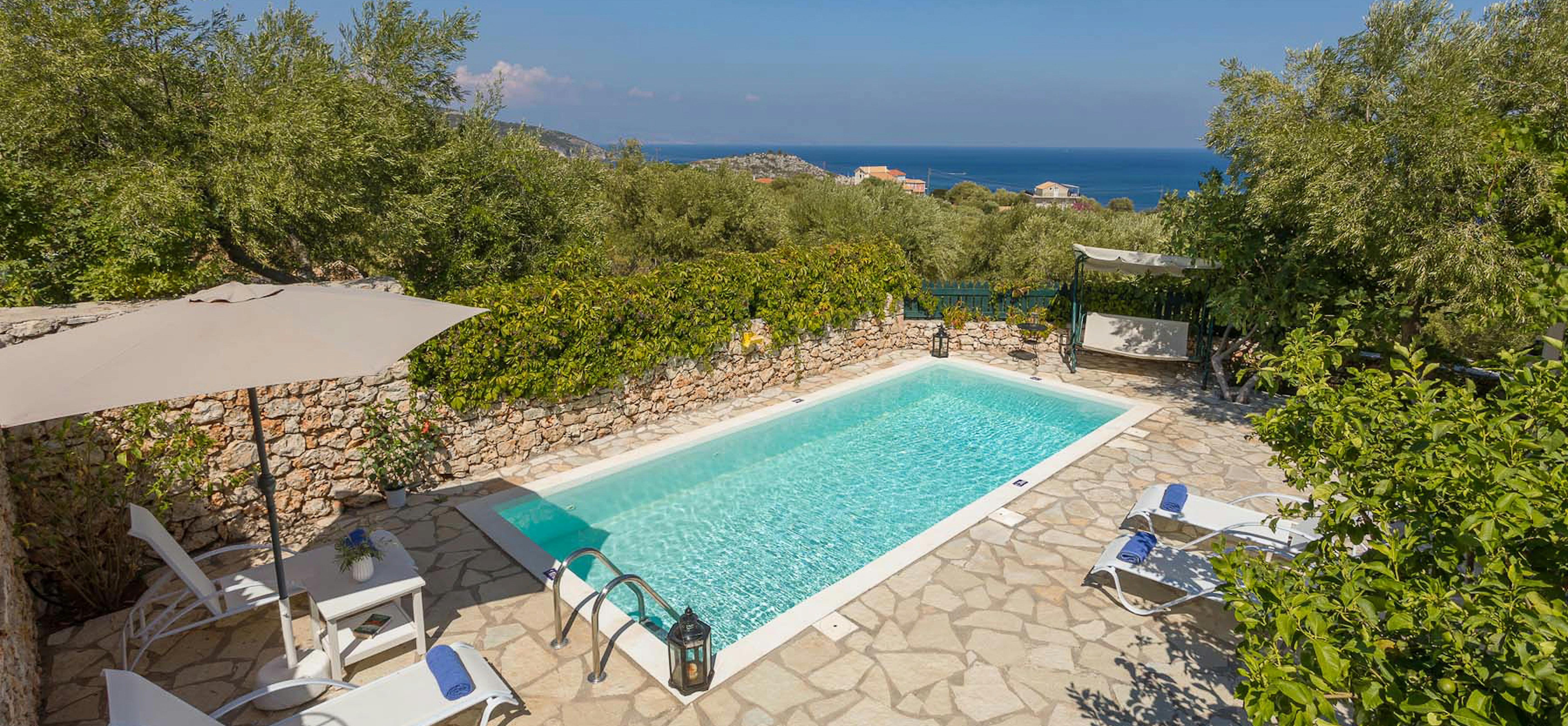 Elevated view of swimming pool with stone terrace, loungers, and coastal sea views framed by olive trees.