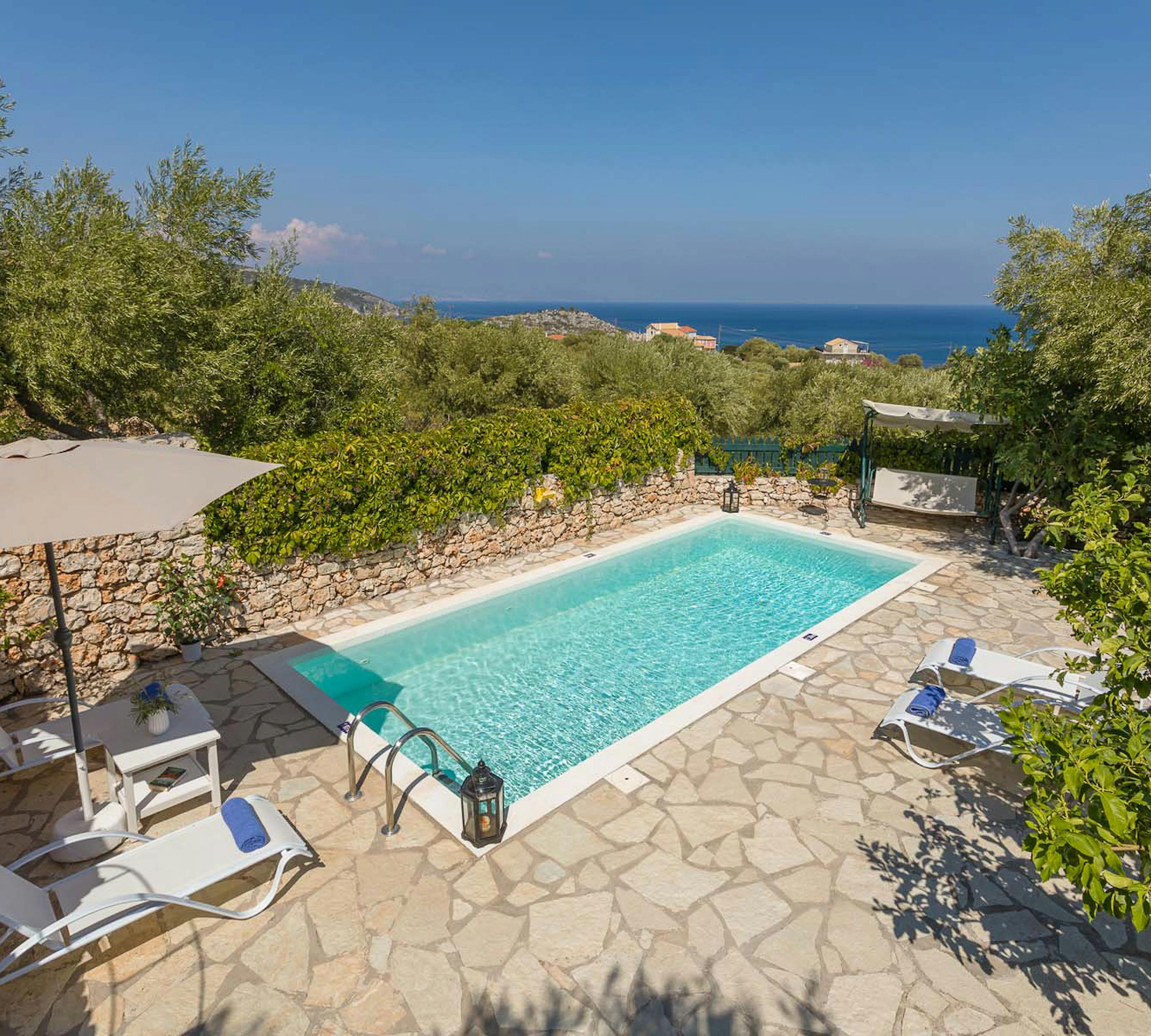 Elevated view of swimming pool with stone terrace, loungers, and coastal sea views framed by olive trees.