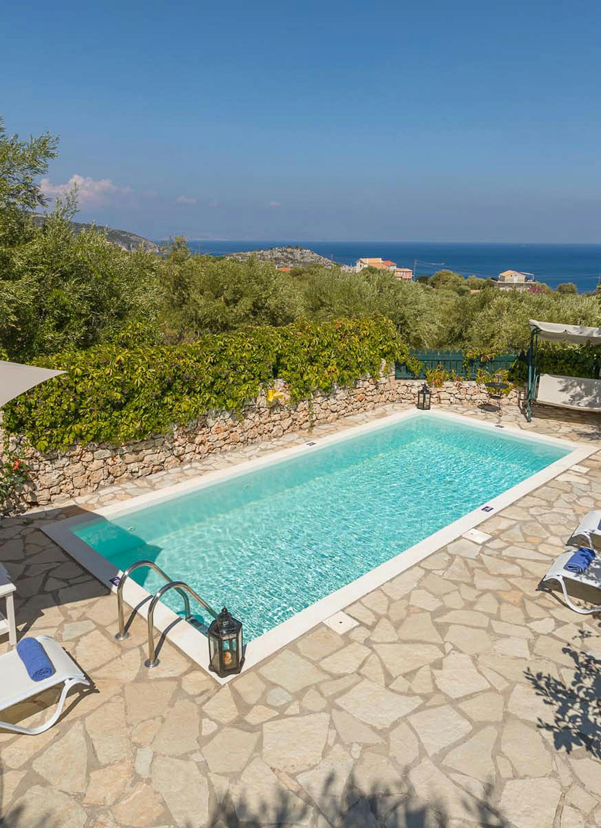 Elevated view of swimming pool with stone terrace, loungers, and coastal sea views framed by olive trees.