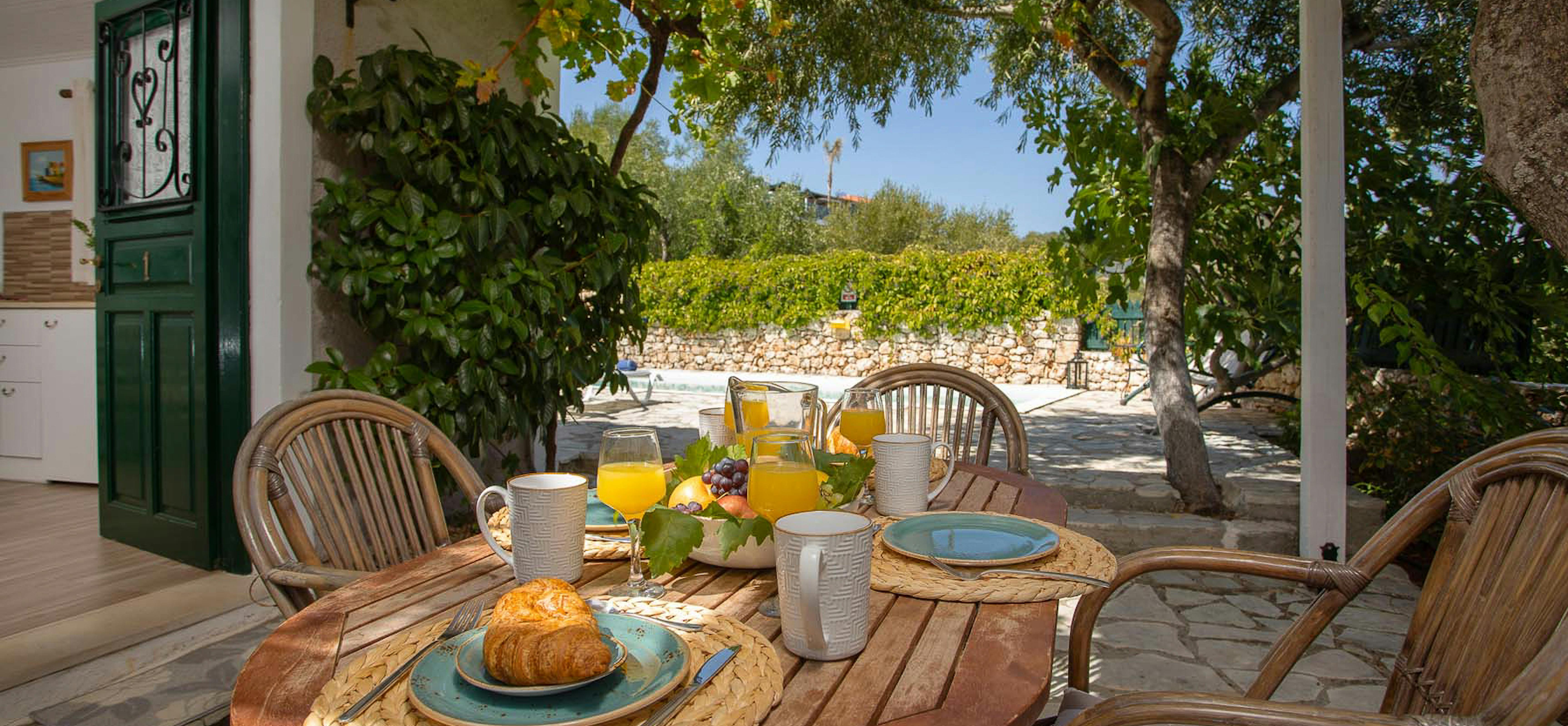 Covered outdoor dining terrace with breakfast setup, wicker chairs, and garden view with stone walls.