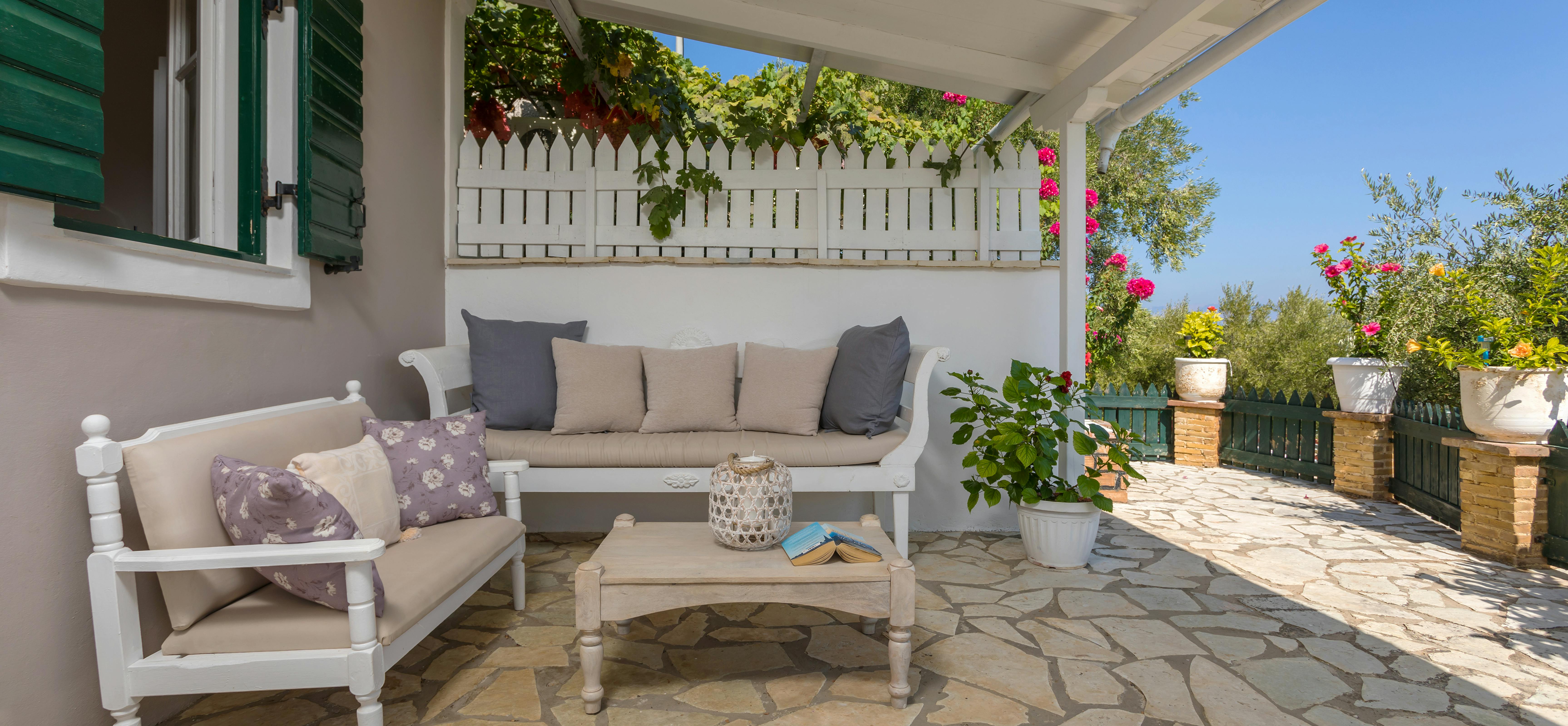Covered terrace with white seating, purple cushions, stone patio, flowering vines, and garden views.