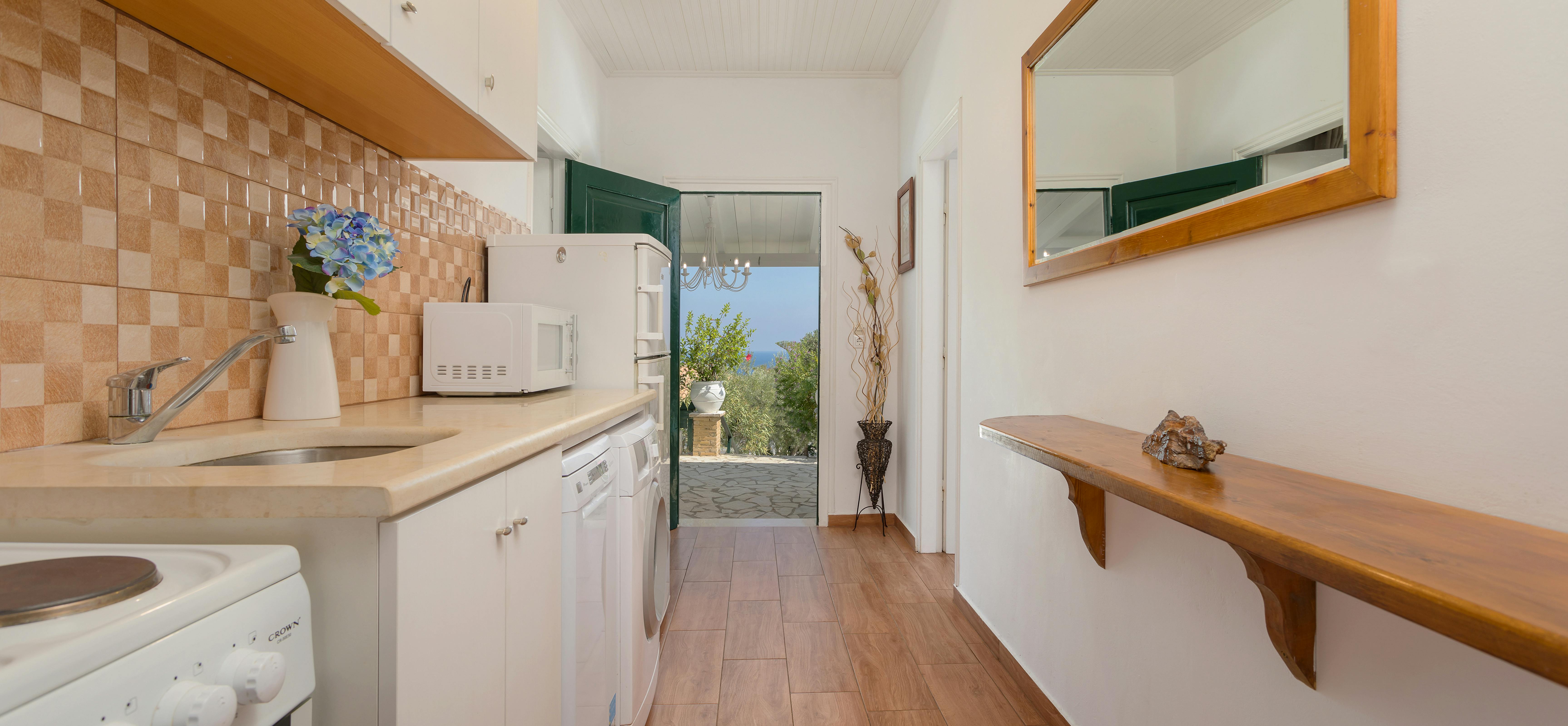 Galley kitchen with beige tile backsplash, white cabinets, wood shelving, and green door to garden.