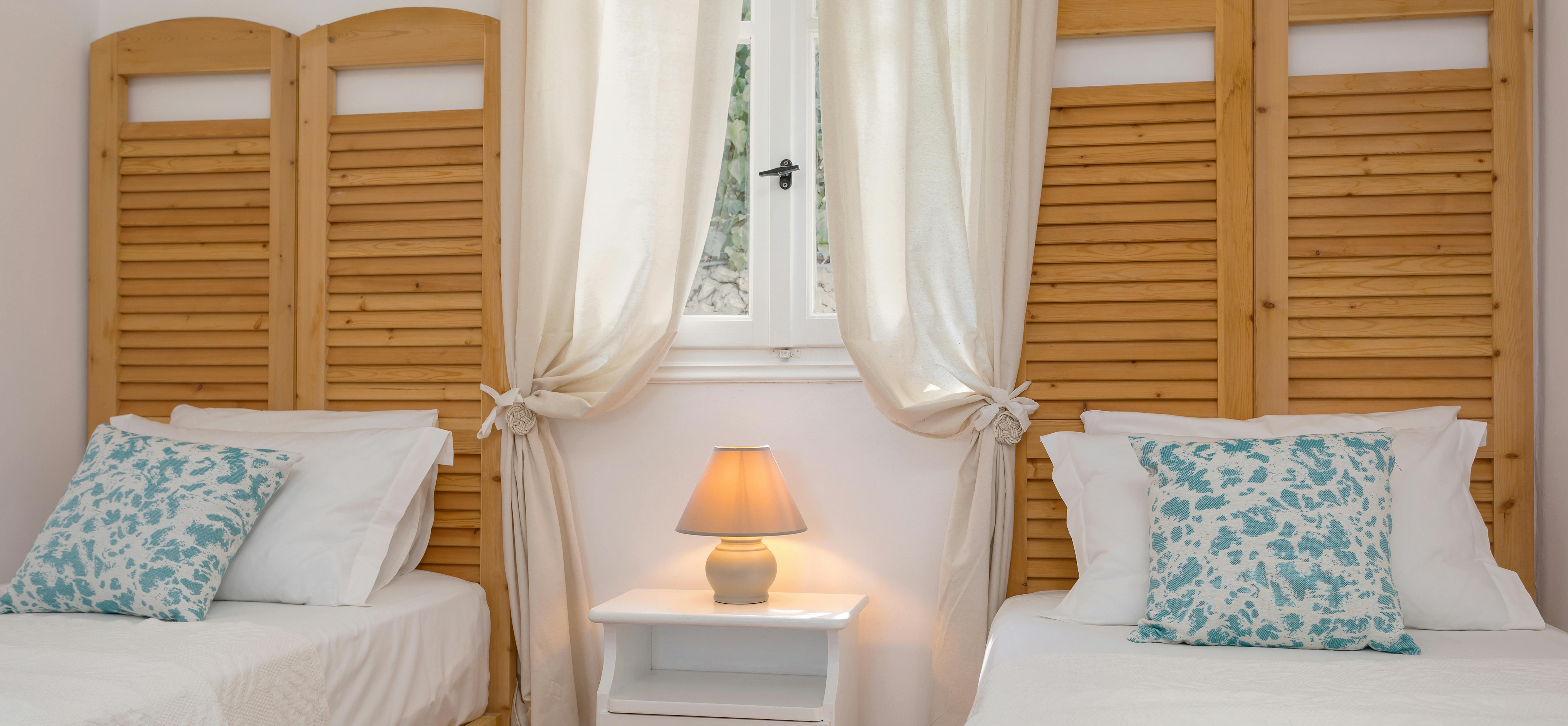 Twin bedroom with wooden shutter headboards, white bedding, turquoise floral cushions, and nightstand.