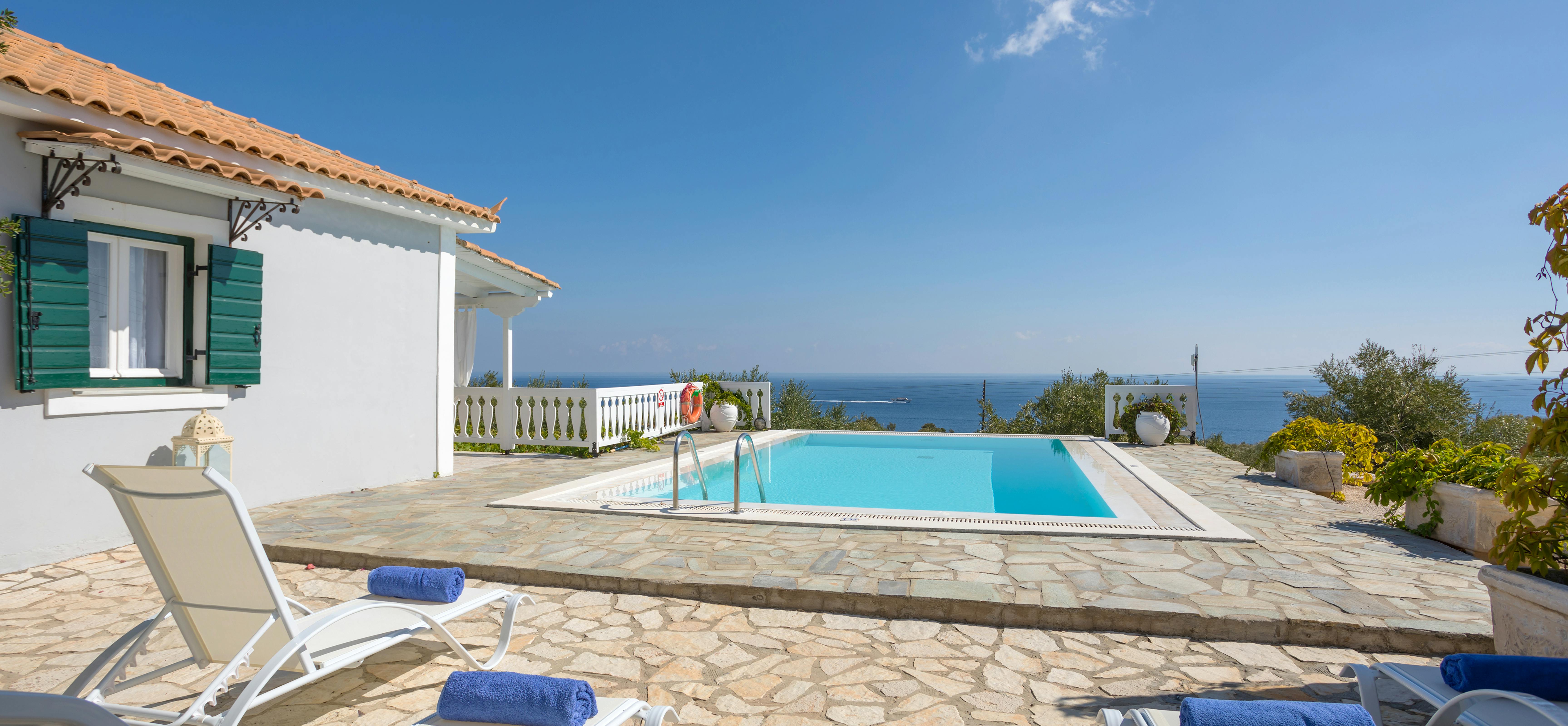 White villa with terracotta roof, swimming pool, sun loungers with blue towels, and coastal sea views.