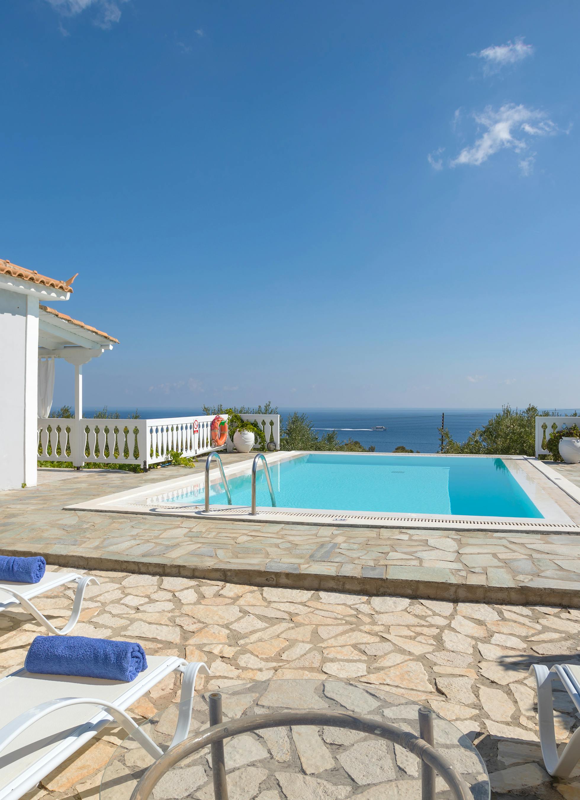 White villa with terracotta roof, swimming pool, sun loungers with blue towels, and coastal sea views.