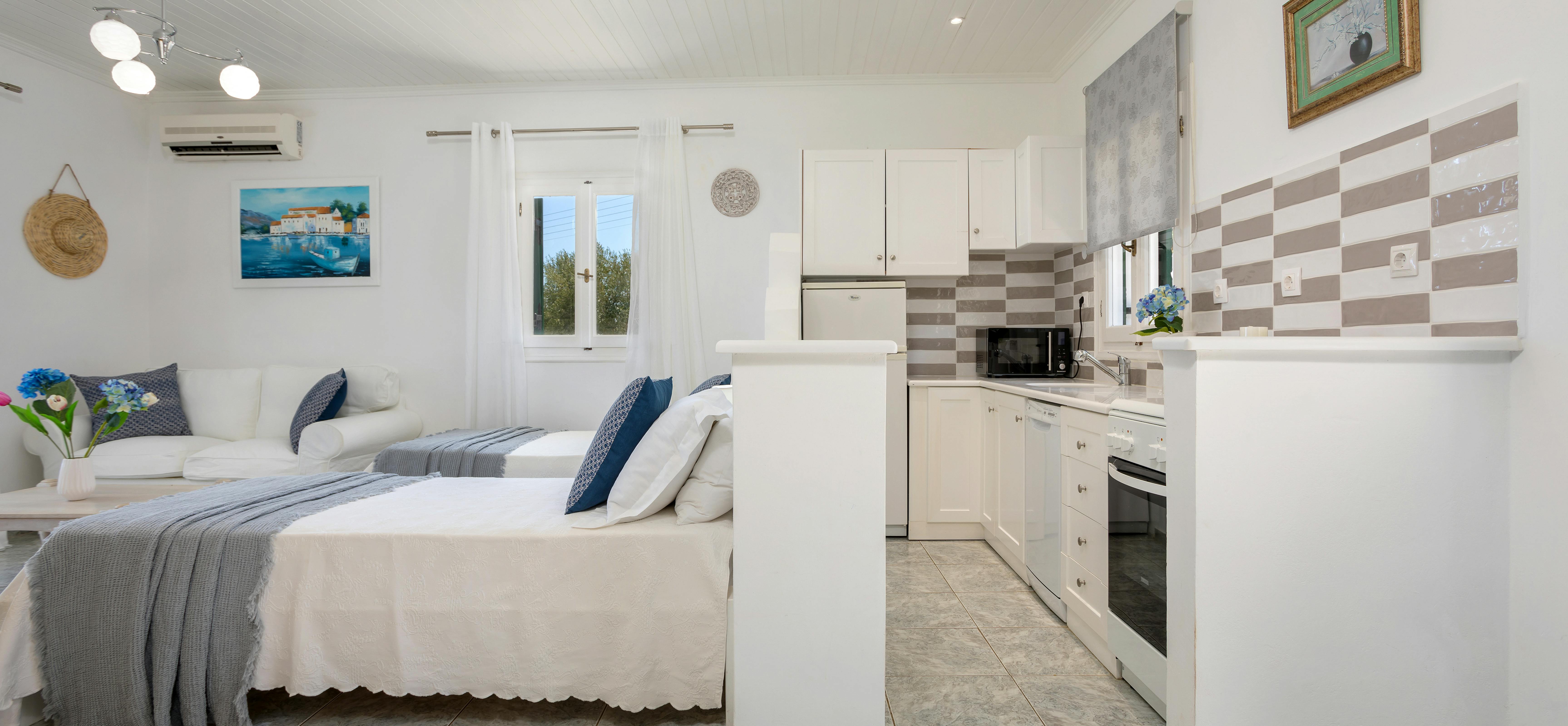 Studio apartment with twin beds, white kitchen, grey throws, and coastal artwork.