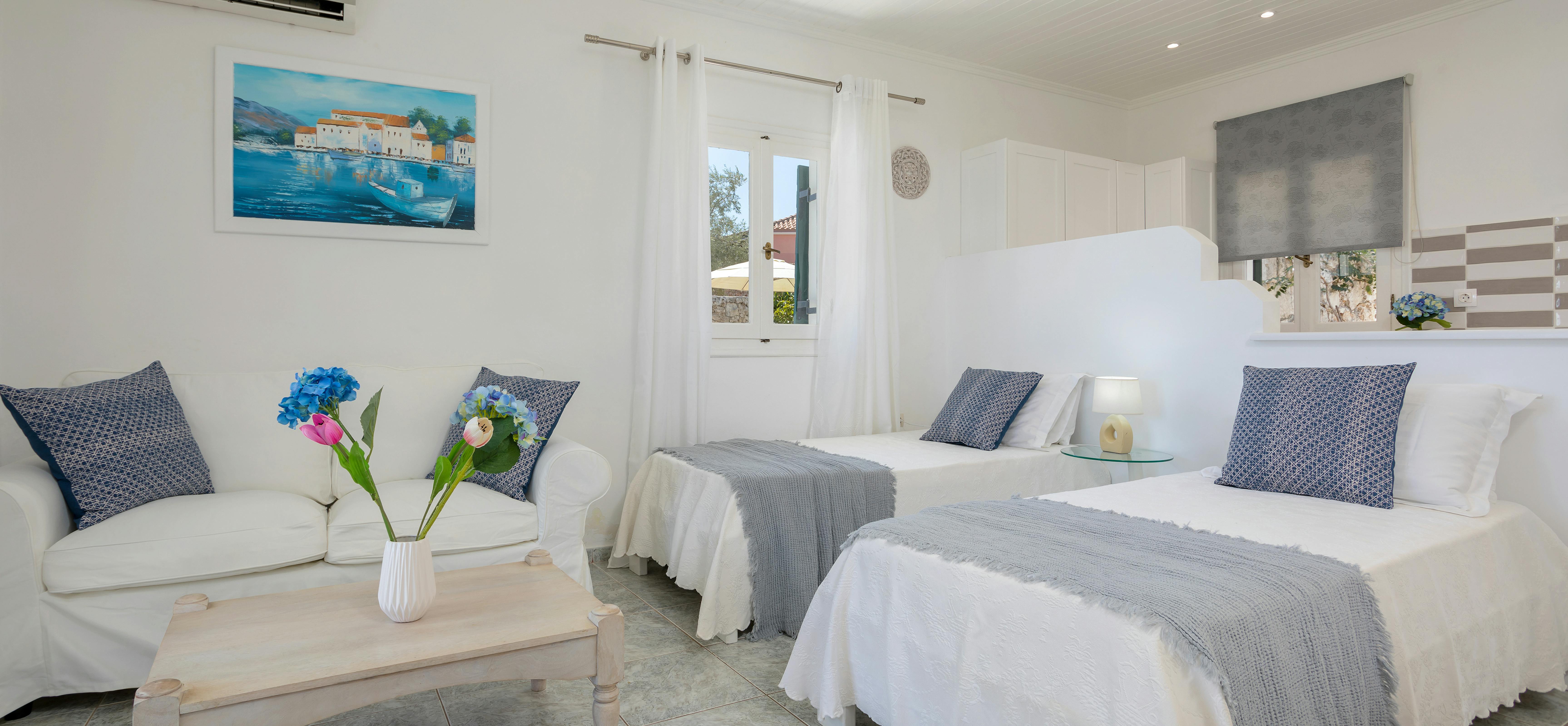 Twin bedroom with white beds, grey throws, blue cushions, seating area, and kitchenette.