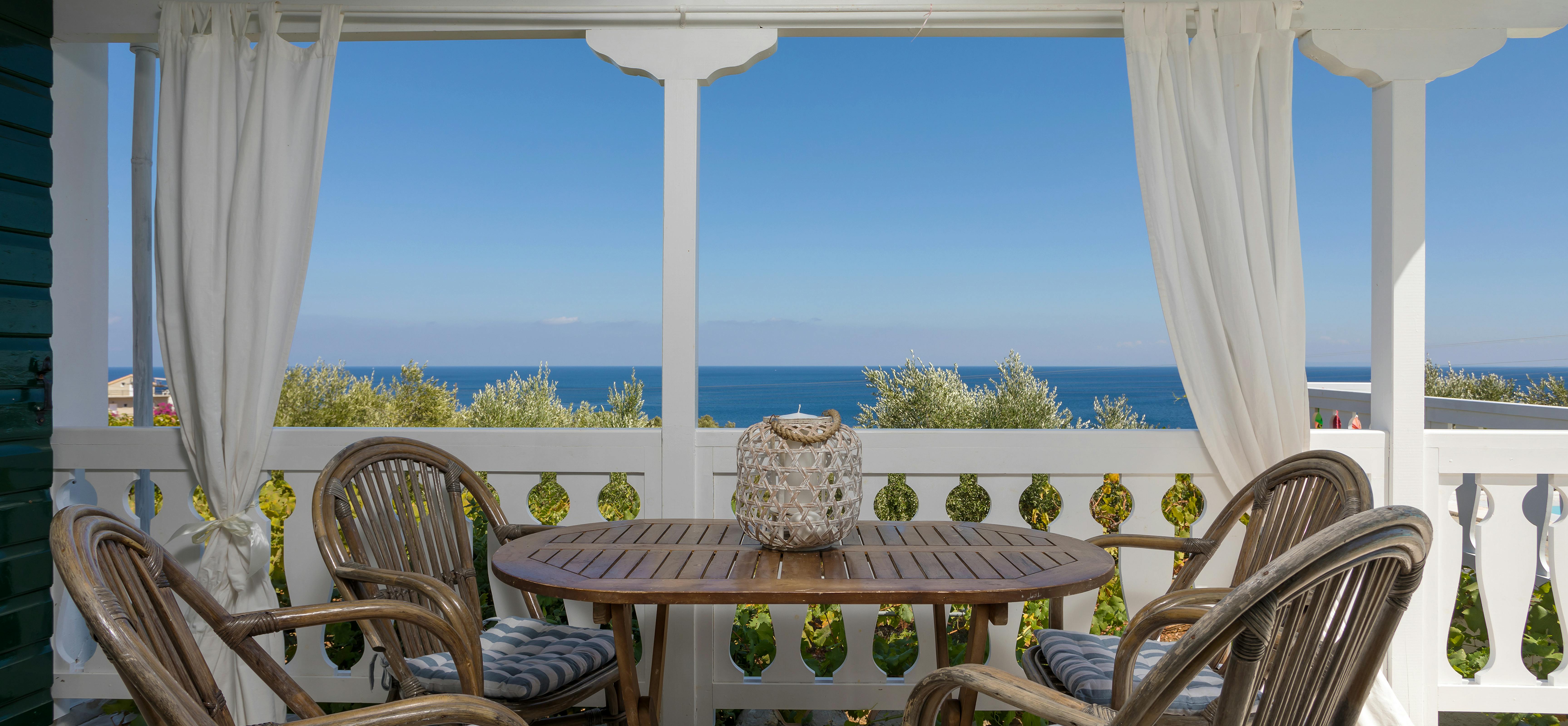 Covered terrace with wicker chairs, striped cushions, wooden table, and open sea horizon views.