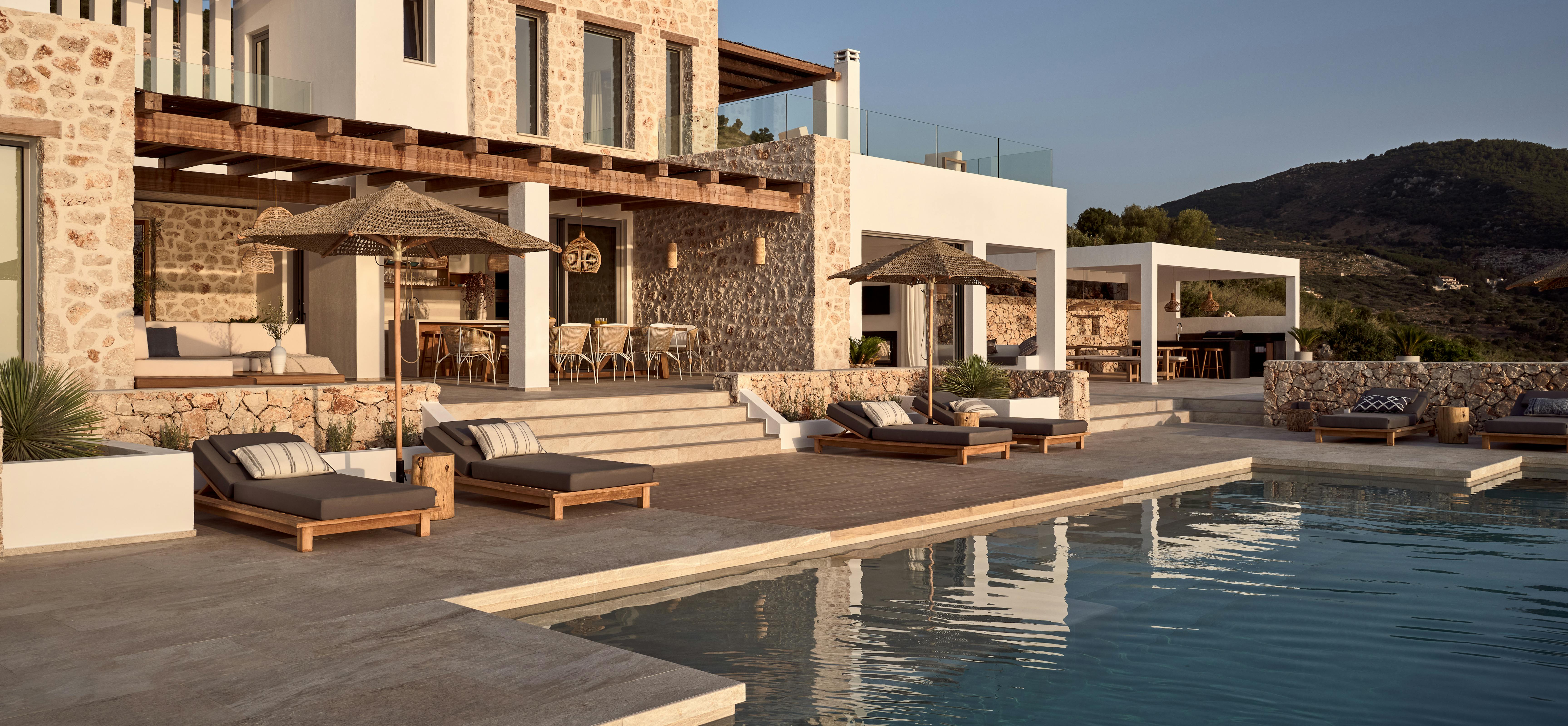 Stone and white villa poolside with wooden sun loungers, woven pendant lights, and panoramic sea views. Multi-level terraces connect covered dining areas with mountain backdrop.