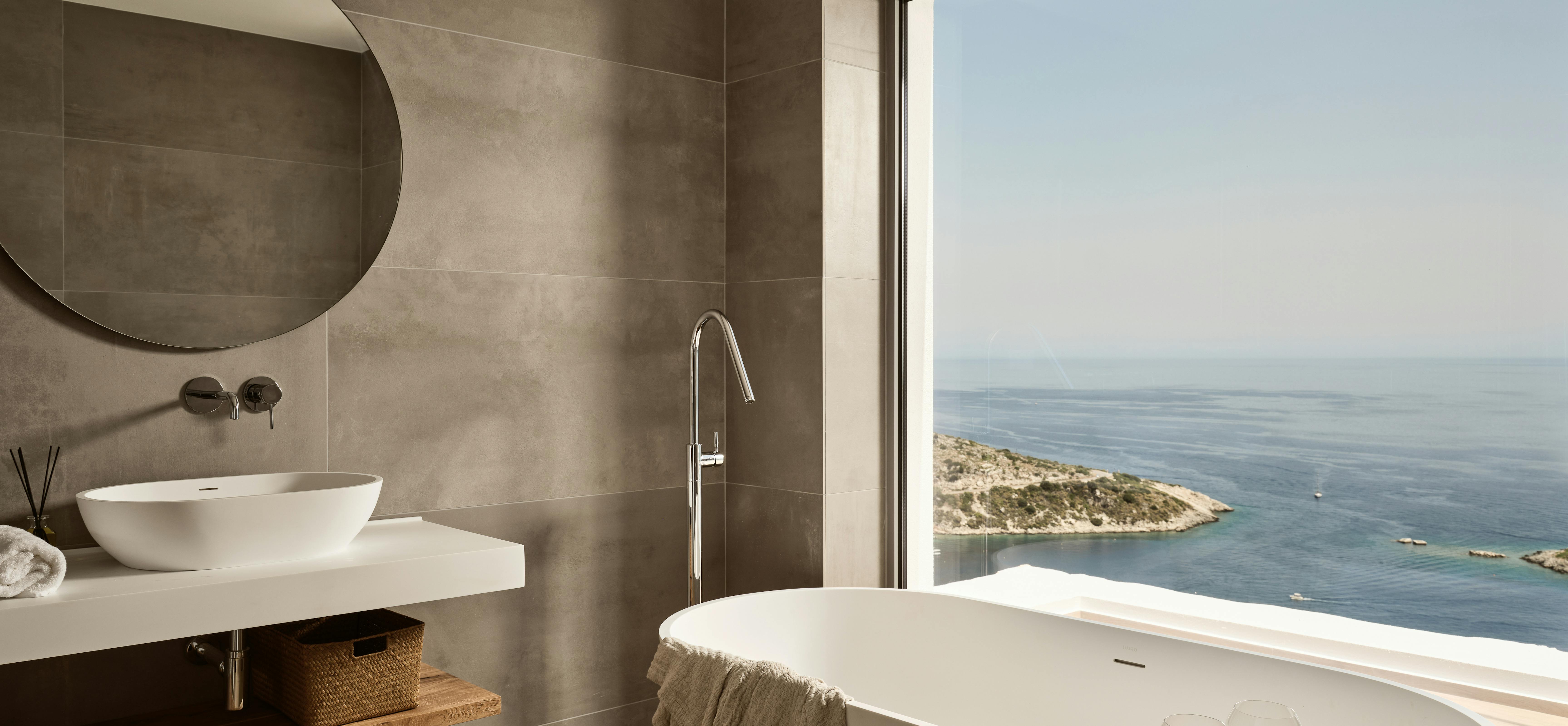 Luxury bathroom with freestanding white tub, vessel sink, and floor-to-ceiling window overlooking coastal waters. Grey concrete tiles and natural wood accents throughout.