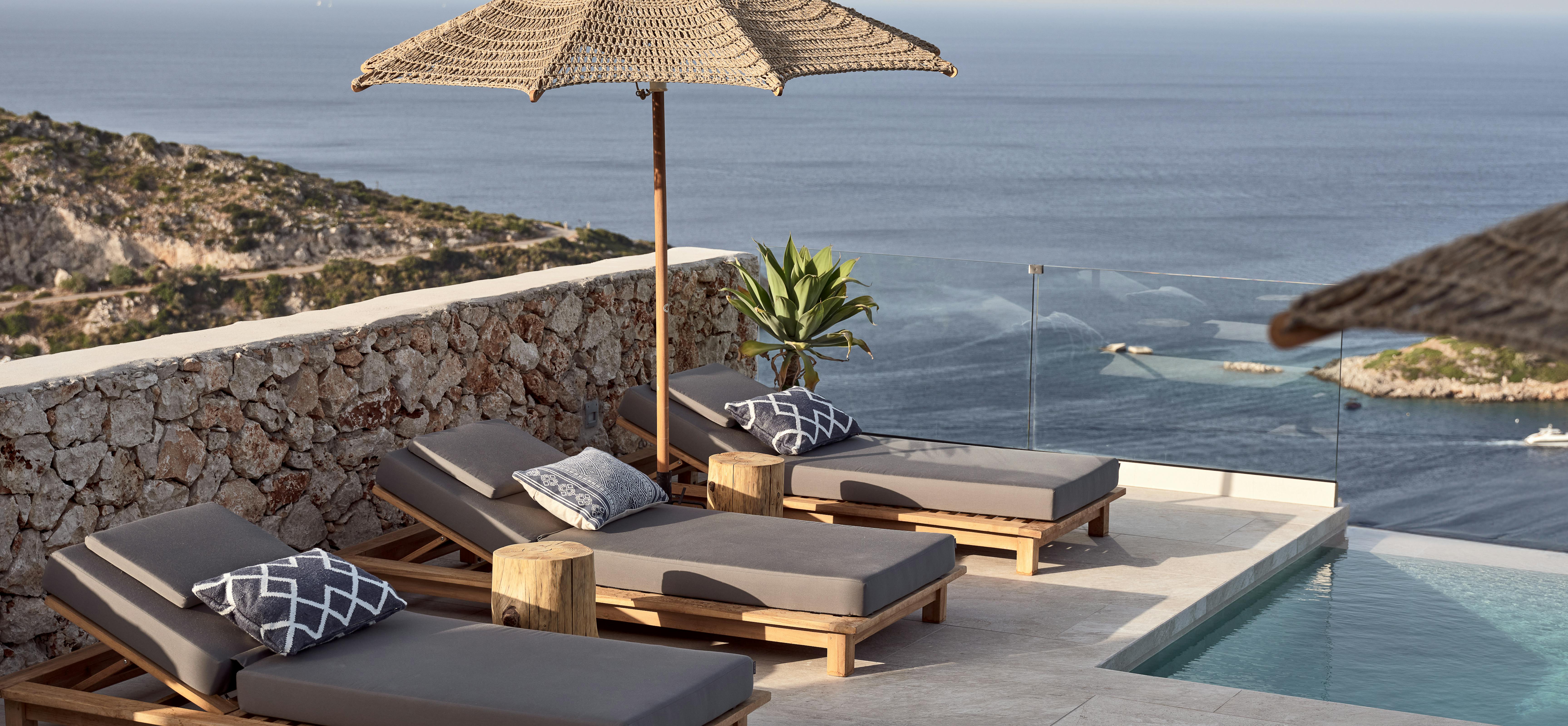 Three wooden loungers with grey cushions and woven umbrella beside infinity pool overlooking dramatic coastal bay. Stone wall and glass railing frame the seascape.
