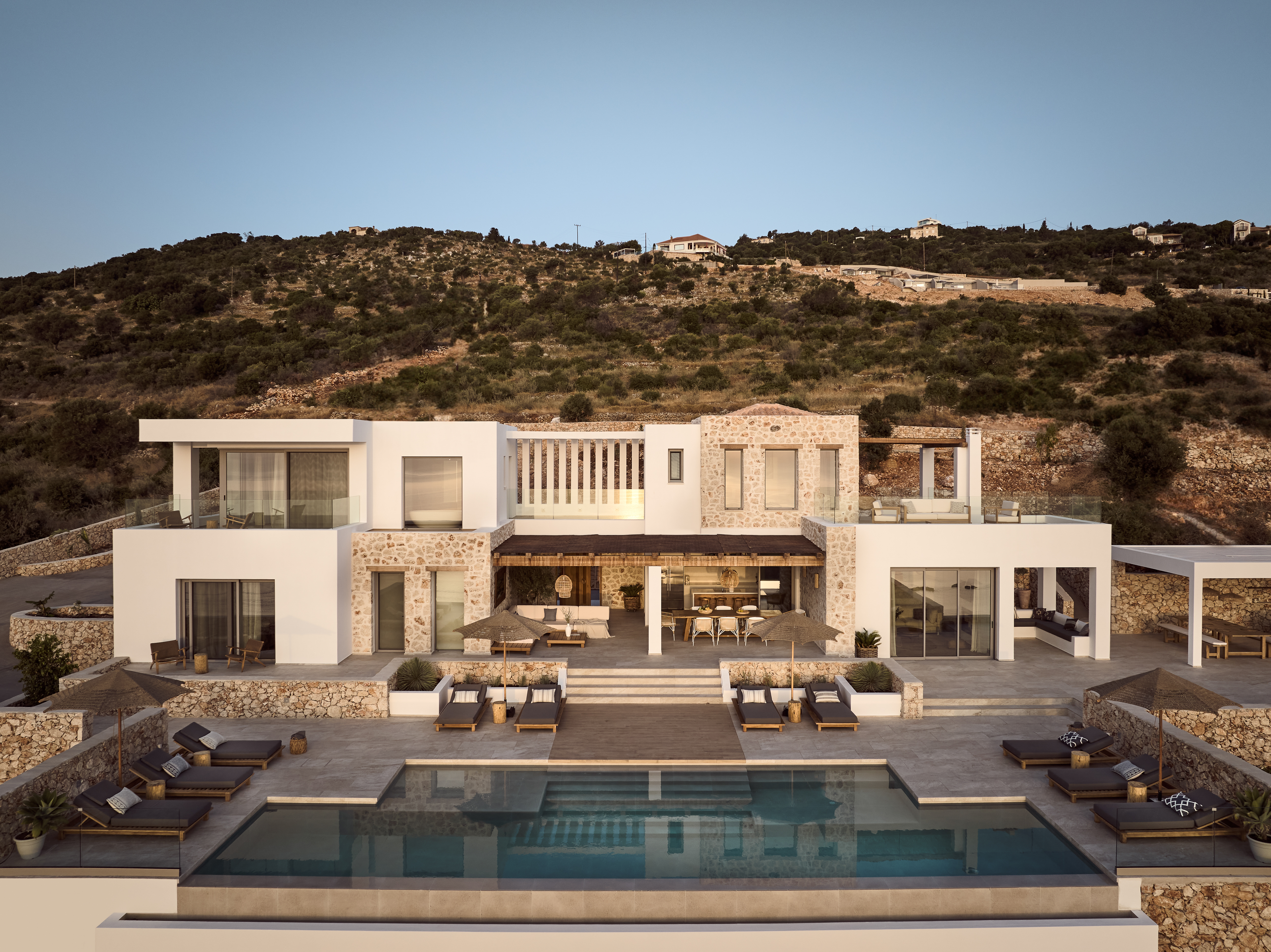 Multi-story Mediterranean villa set into hillside with swimming pool, terraced lounging areas, and wooden sun beds. White and stone architecture with expansive outdoor living spaces.