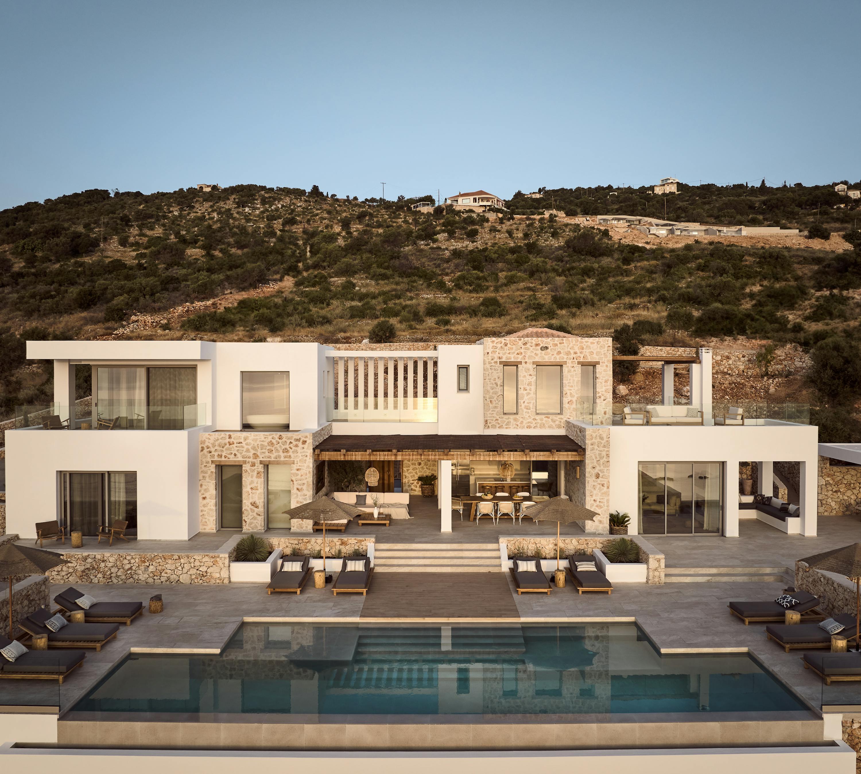 Multi-story Mediterranean villa set into hillside with swimming pool, terraced lounging areas, and wooden sun beds. White and stone architecture with expansive outdoor living spaces.