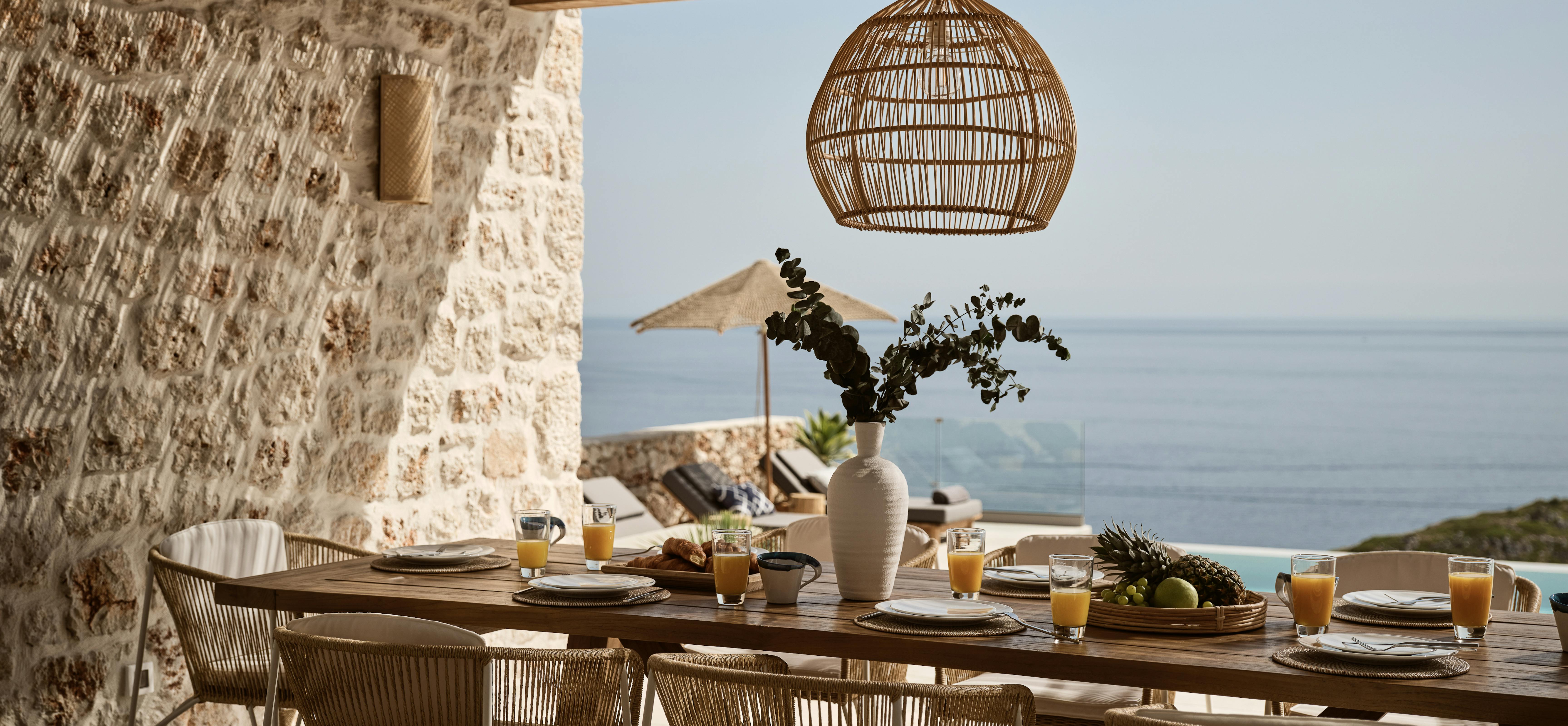 Outdoor dining table set for breakfast under pergola with ocean views and loungers visible beyond. Woven pendant light and stone walls complement the coastal setting.