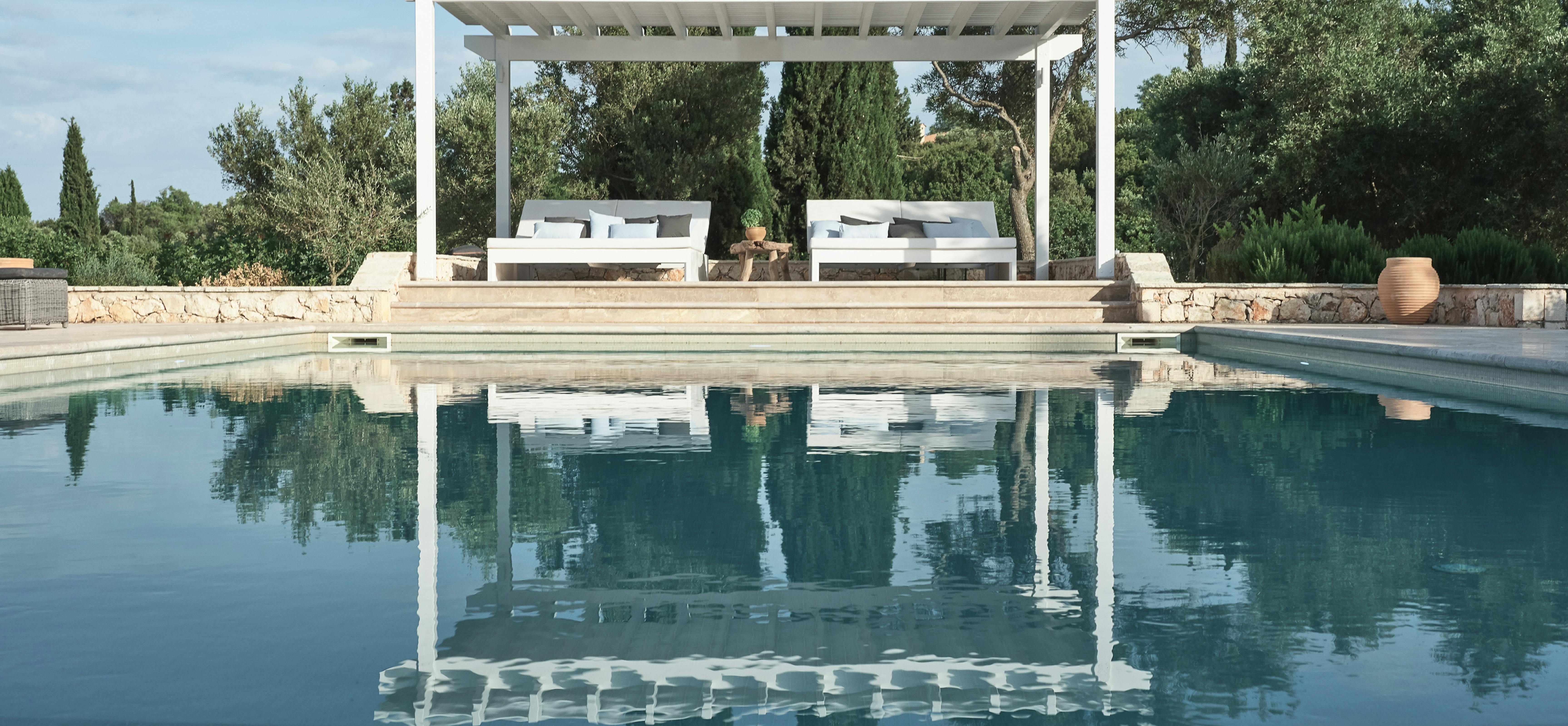 Infinity pool reflecting white pergola with daybeds, backed by cypress trees and Mediterranean landscape.
