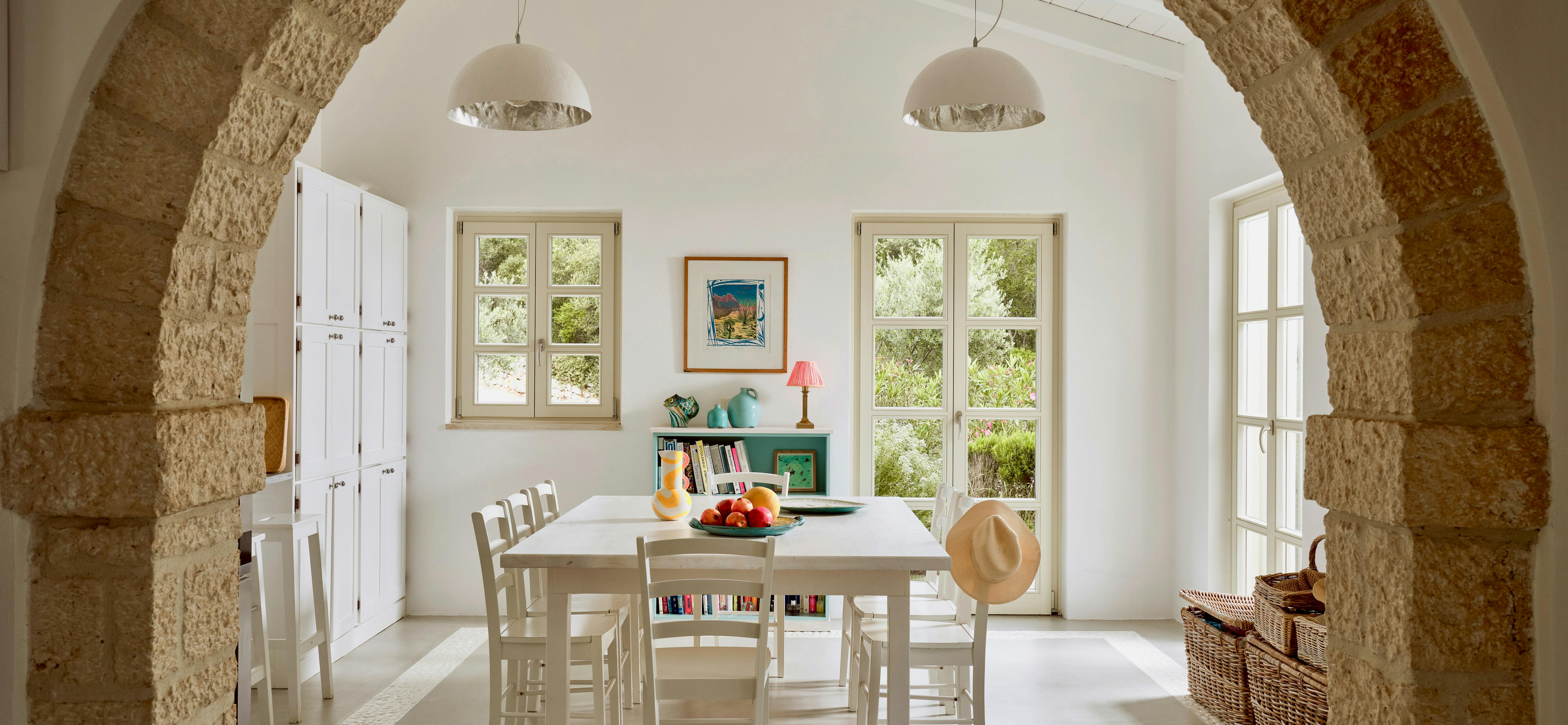 White dining area framed by rustic stone arch with pendant lights overhead. Simple white table and chairs surrounded by tall windows overlooking garden views.