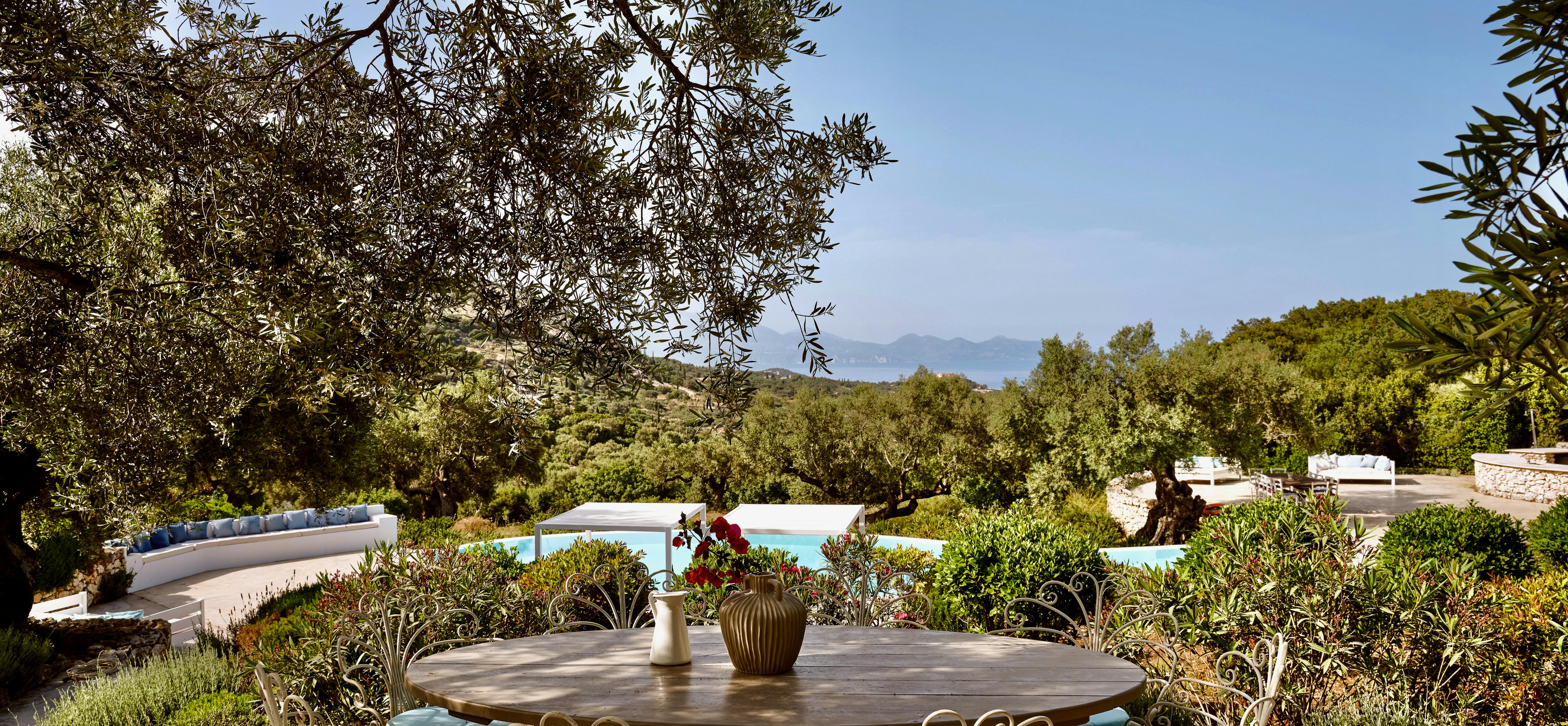 Garden dining area with vintage metal chairs around round table beneath olive trees. Pool and coastal views create idyllic outdoor setting.