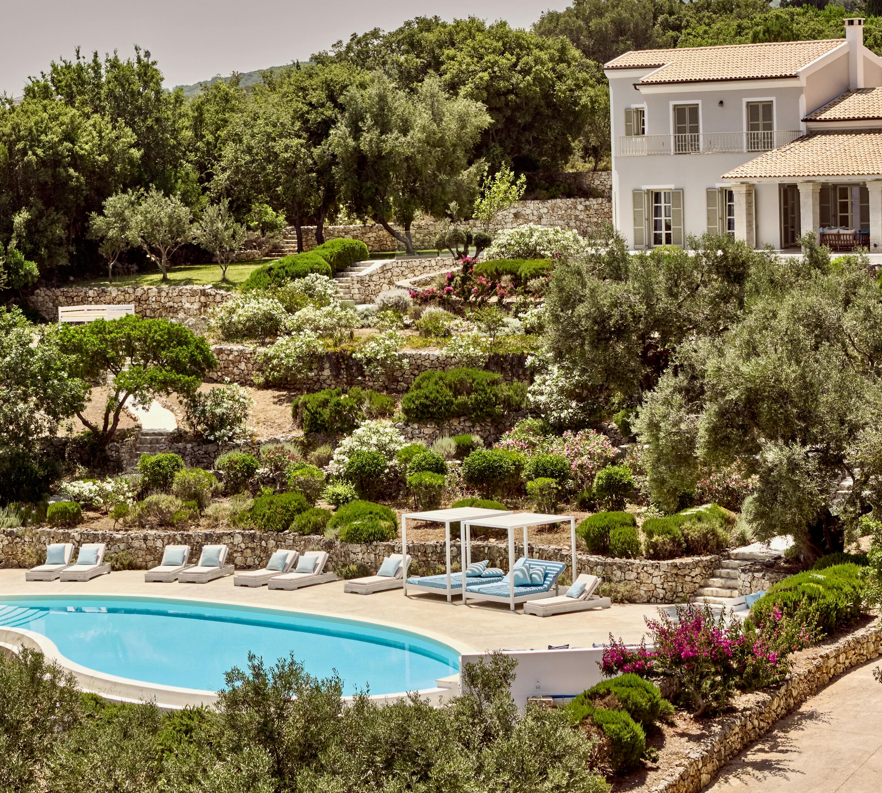 Hillside estate with white villa overlooking terraced gardens and curved pool below. Stone retaining walls and mature olive trees frame the Mediterranean landscape setting.