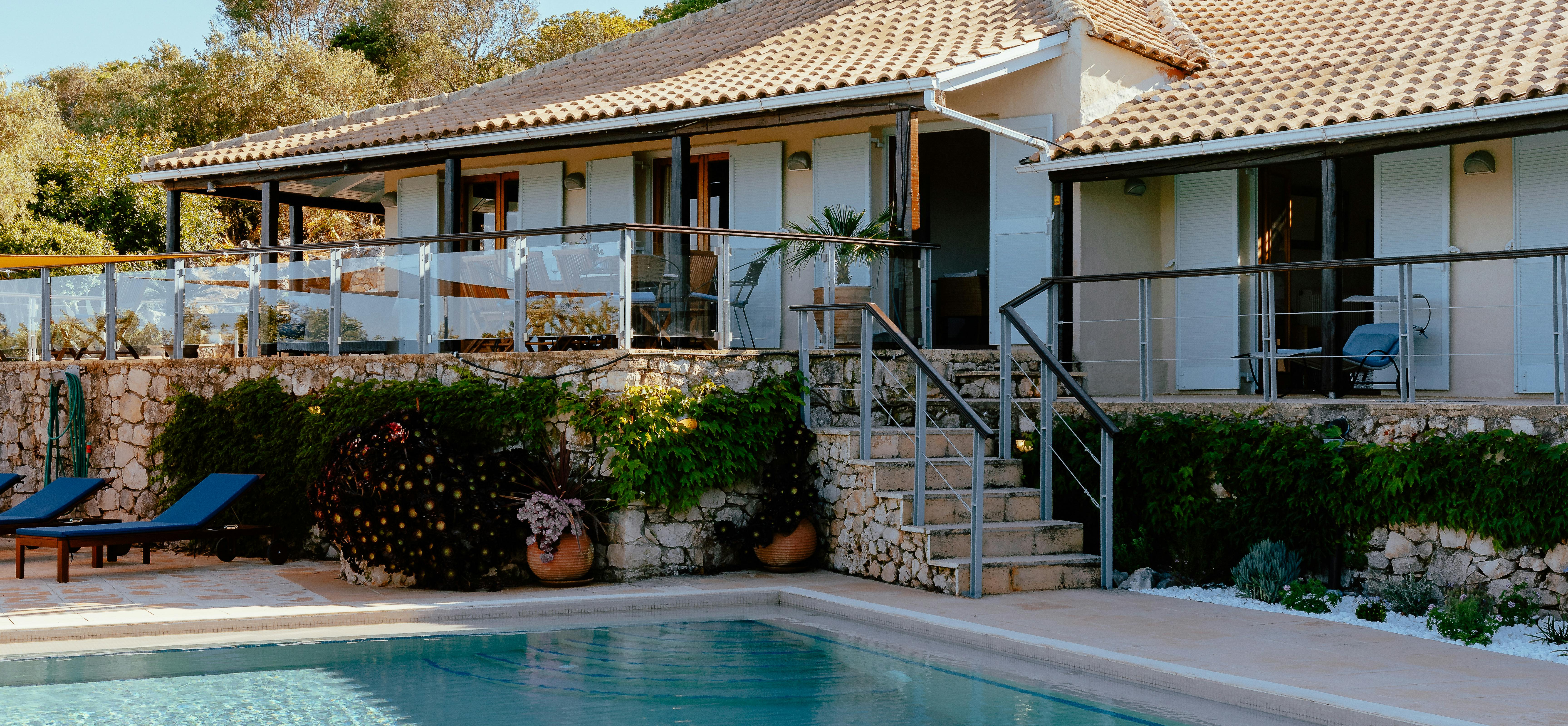 The villa's white facade with terracotta roof and turret feature rises above the sparkling pool, with stone terraces connected by modern glass-railed stairs.
