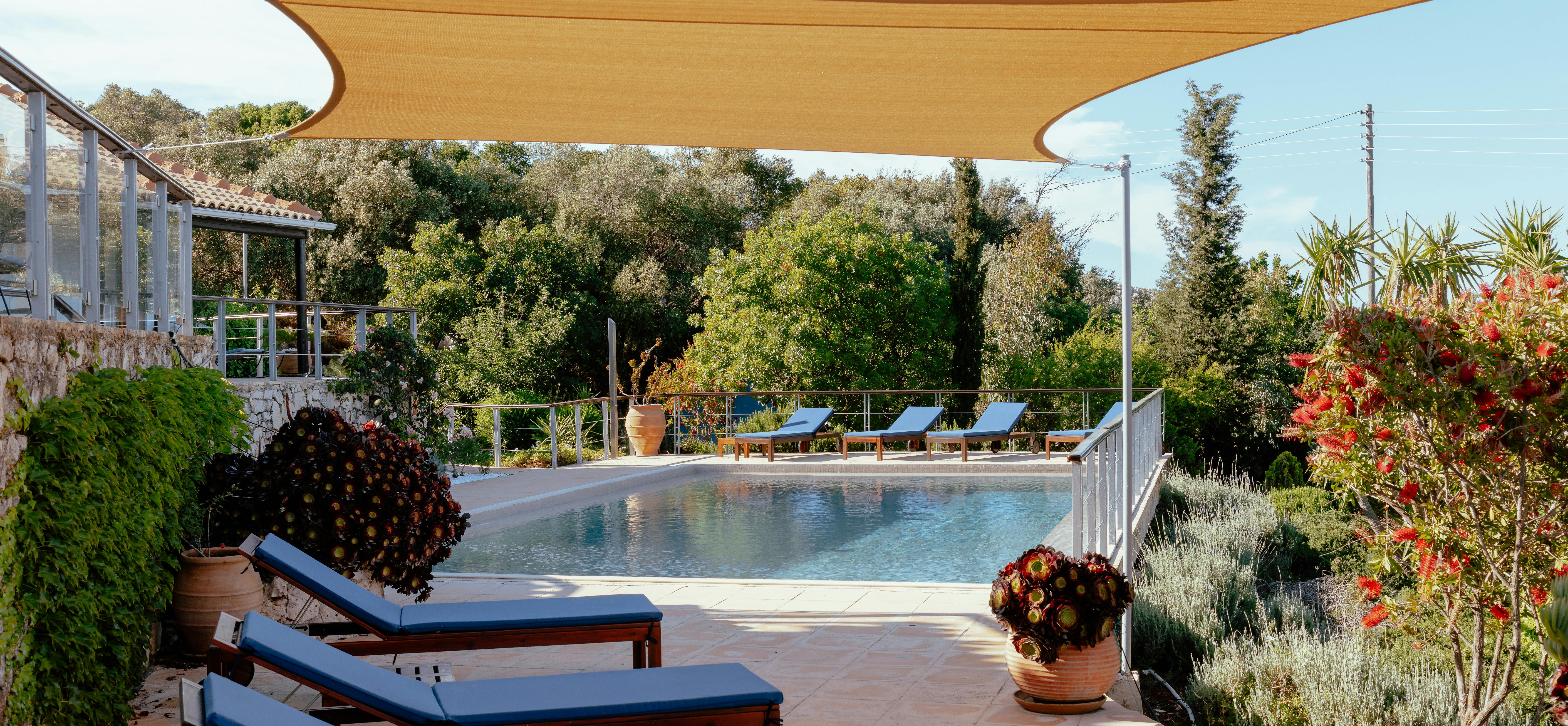 A poolside terrace shaded by a large golden fabric sail features blue-cushioned loungers, Mediterranean gardens with herbs and flowering plants, and white metal railings.
