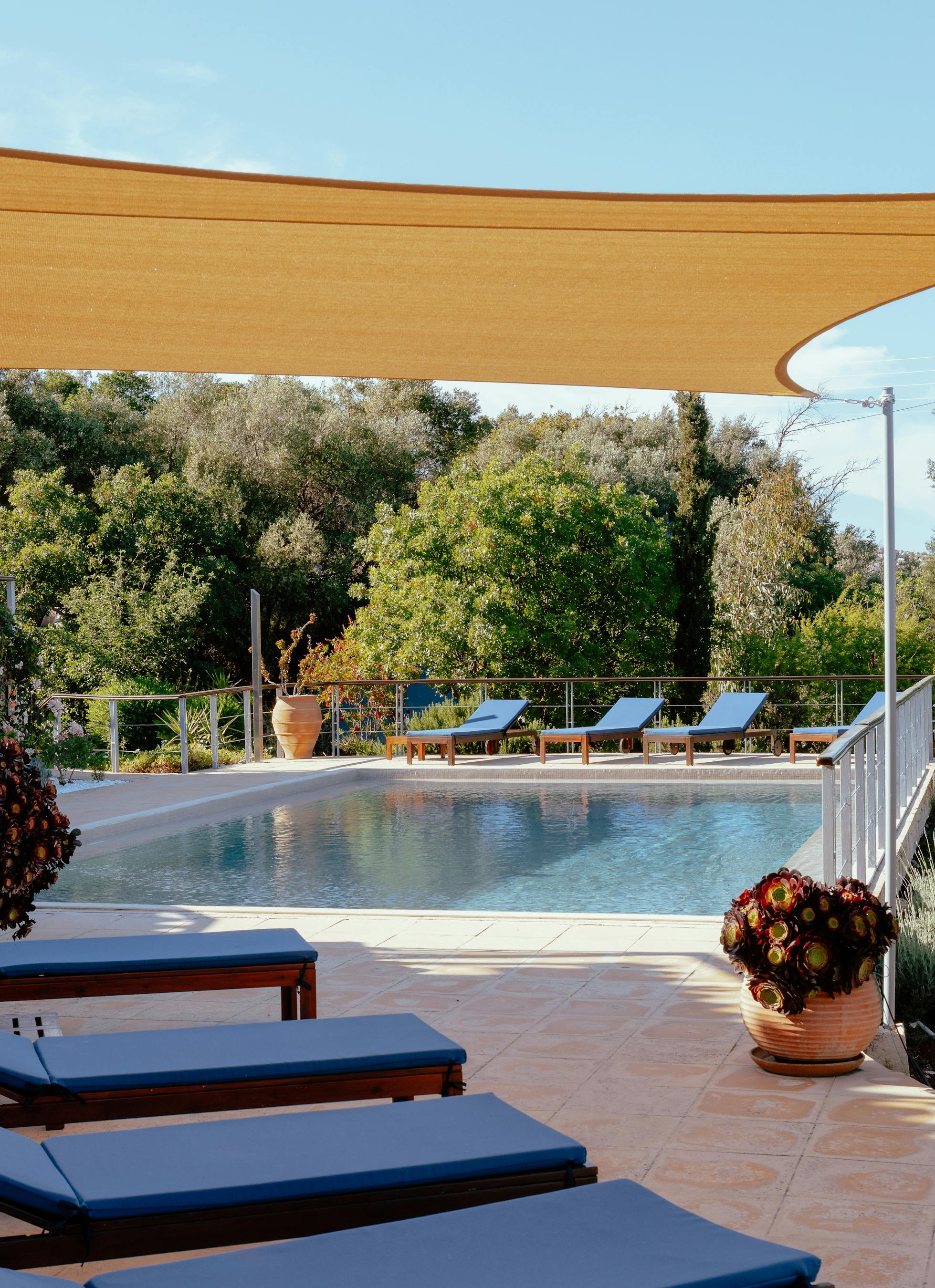 A poolside terrace shaded by a large golden fabric sail features blue-cushioned loungers, Mediterranean gardens with herbs and flowering plants, and white metal railings.