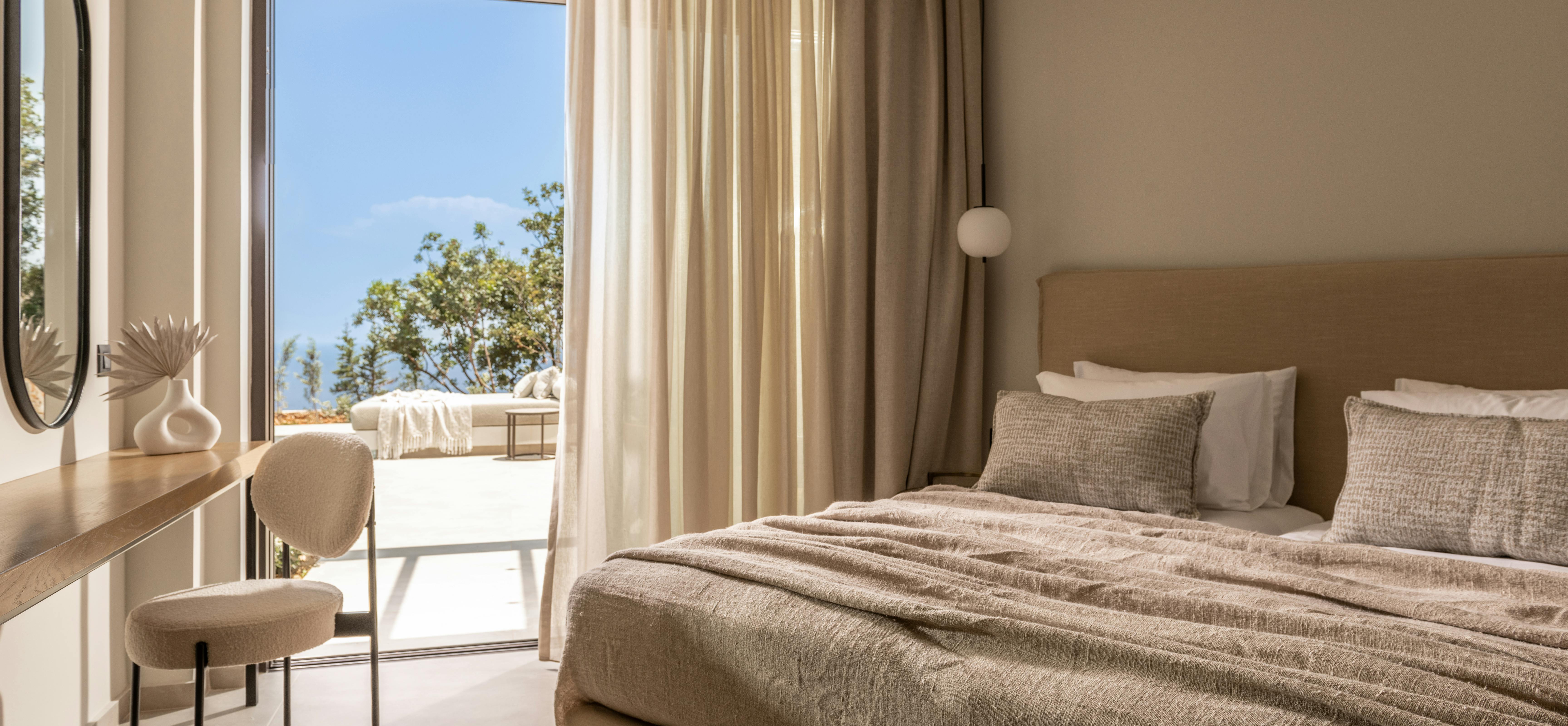 A minimalist bedroom opens to an ocean vista through glass doors with flowing curtains, featuring a plush bed dressed in textured beige linens.