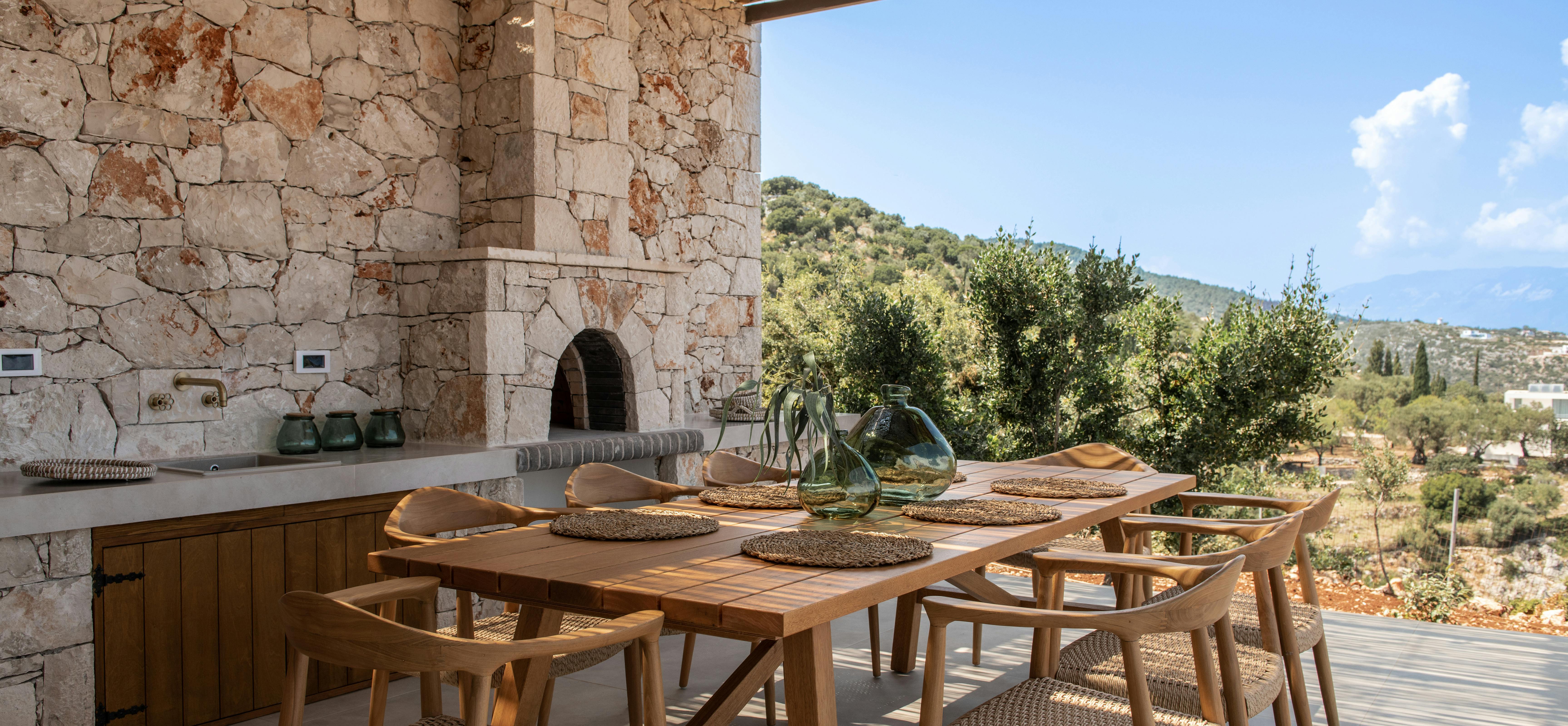 A covered outdoor dining area incorporates weathered stone walls, a wooden table with woven chairs, and views of the surrounding olive groves.
