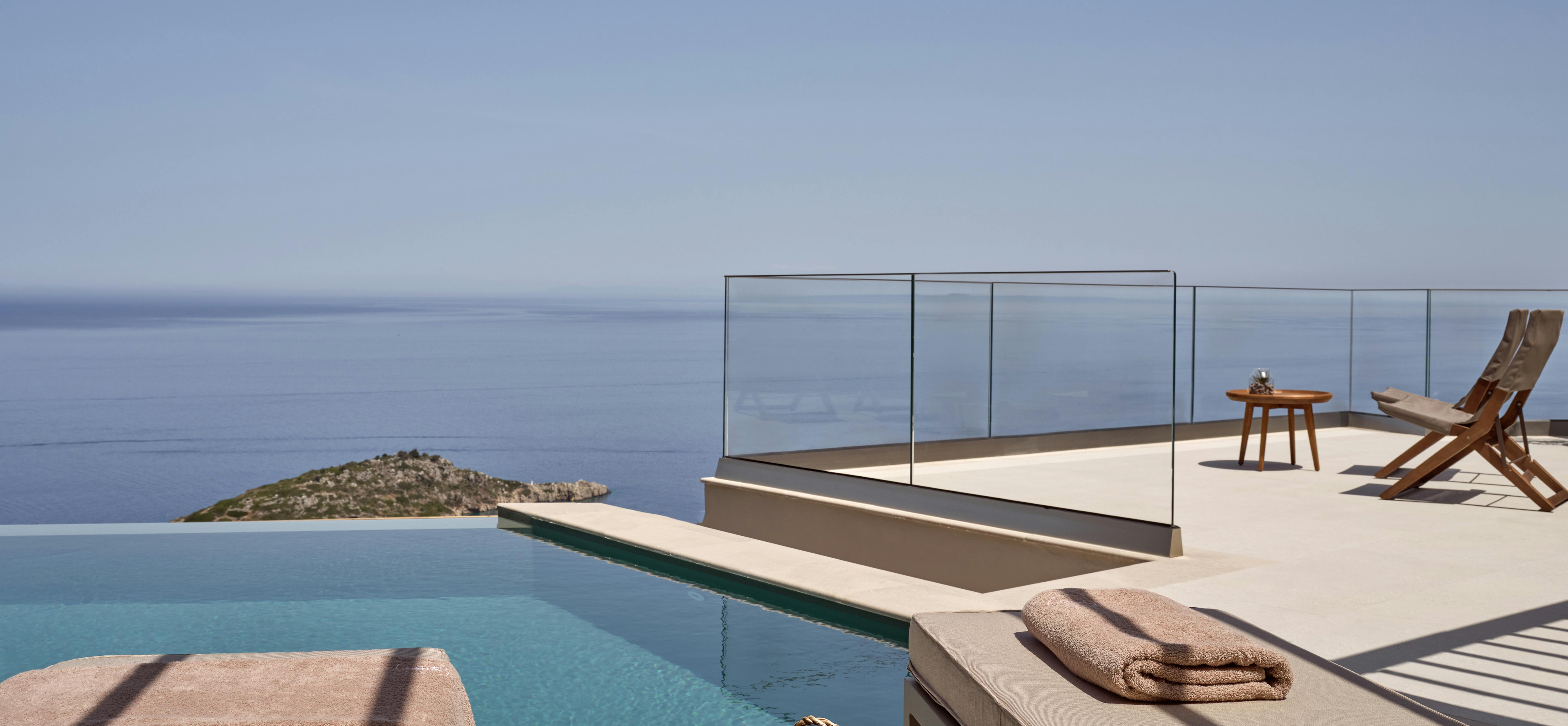 A poolside deck overlooking the Mediterranean Sea with modern sun loungers, a woven basket, and frameless glass railings.