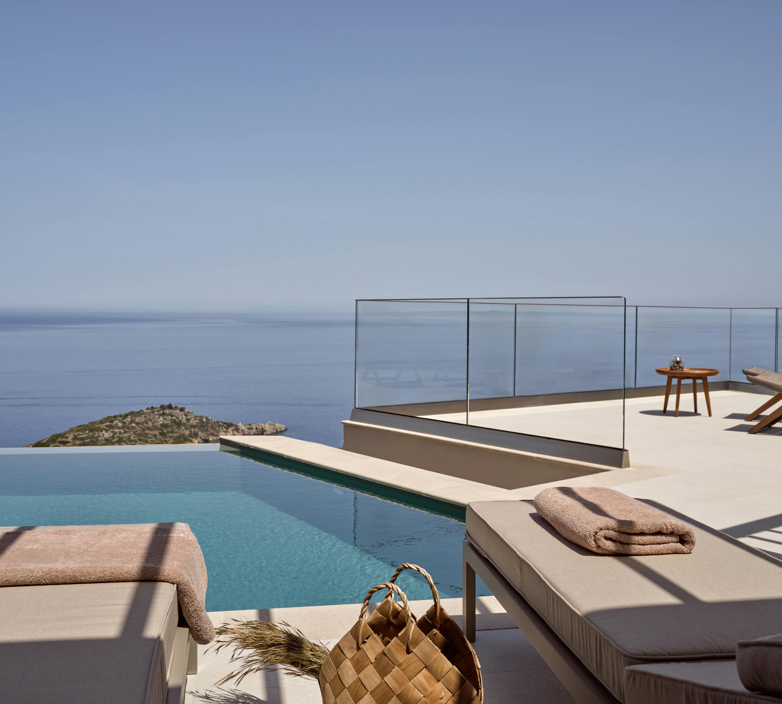 A poolside deck overlooking the Mediterranean Sea with modern sun loungers, a woven basket, and frameless glass railings.