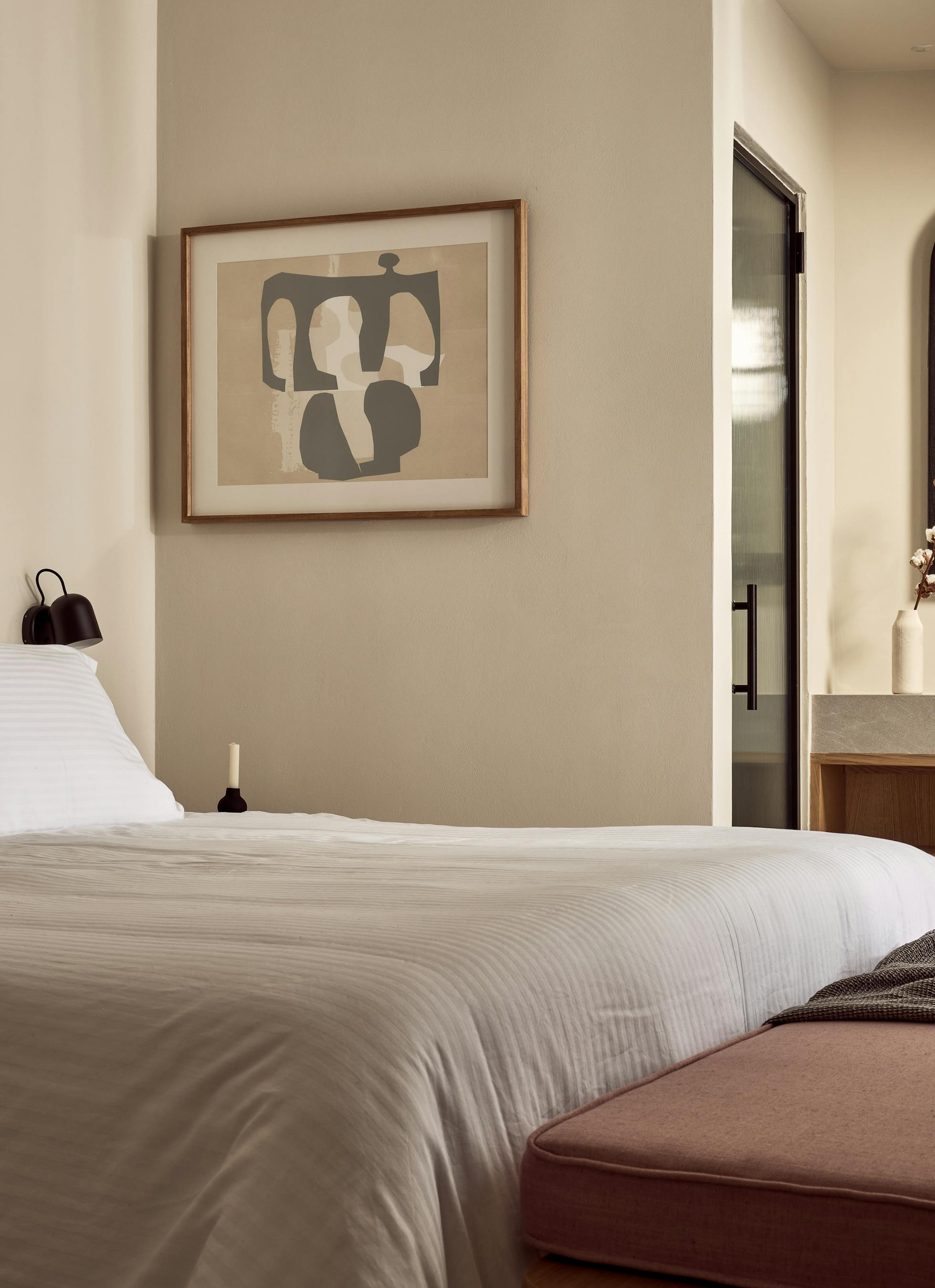 A serene bedroom with cane-backed headboard, abstract artwork, white bedding with mauve accents, and arched doorway to an ensuite bathroom.