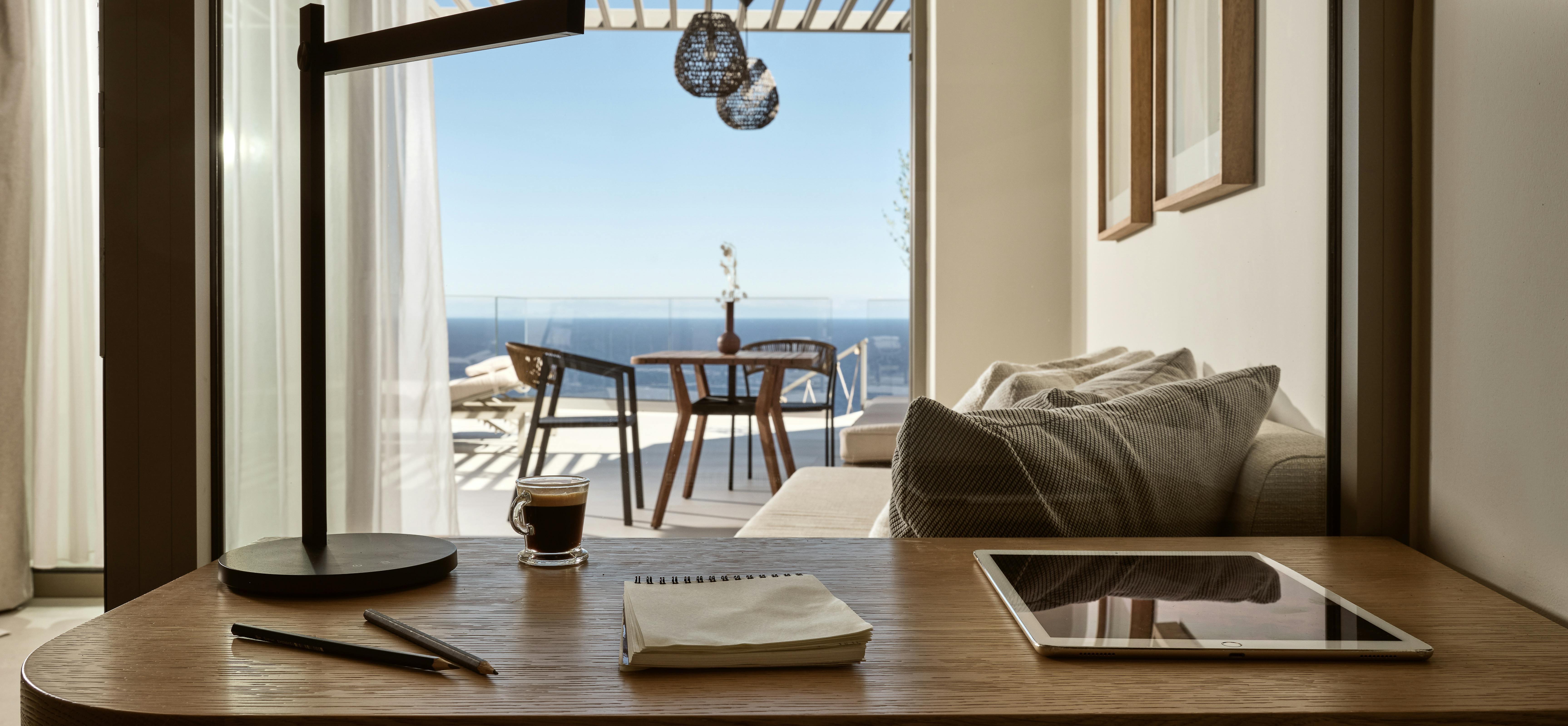 A workspace with a curved wooden desk, modern task lamp, and ocean views through floor-to-ceiling windows leading to a terrace with outdoor seating.