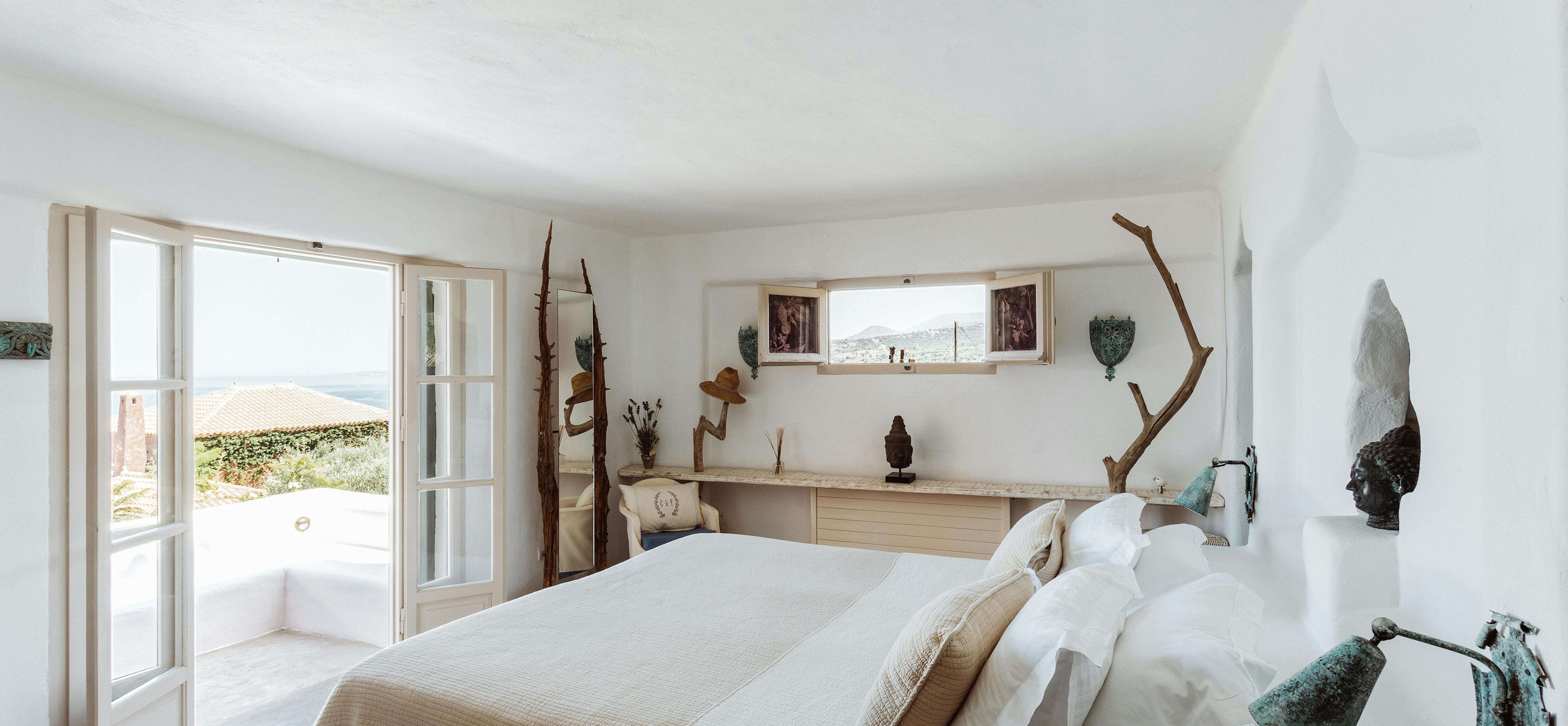 A serene white bedroom with French doors opening to a terrace, driftwood branches as wall art, rustic wood floating shelf with decorative objects, white bedding with beige throw, weathered bronze sculptures, and ocean views.