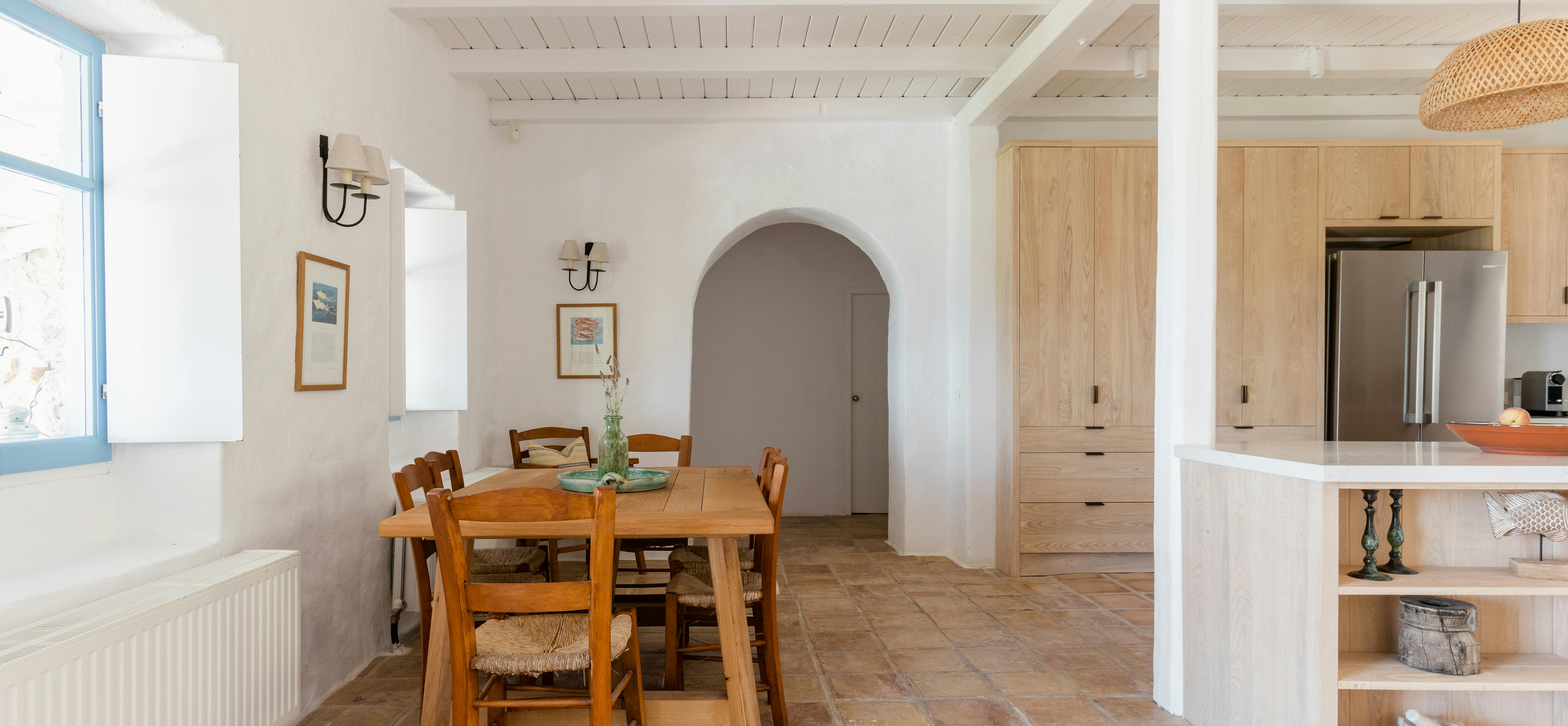 An open-plan Mediterranean dining area with rustic wood table, arched doorway, light oak kitchen cabinetry, and terracotta tile floor.