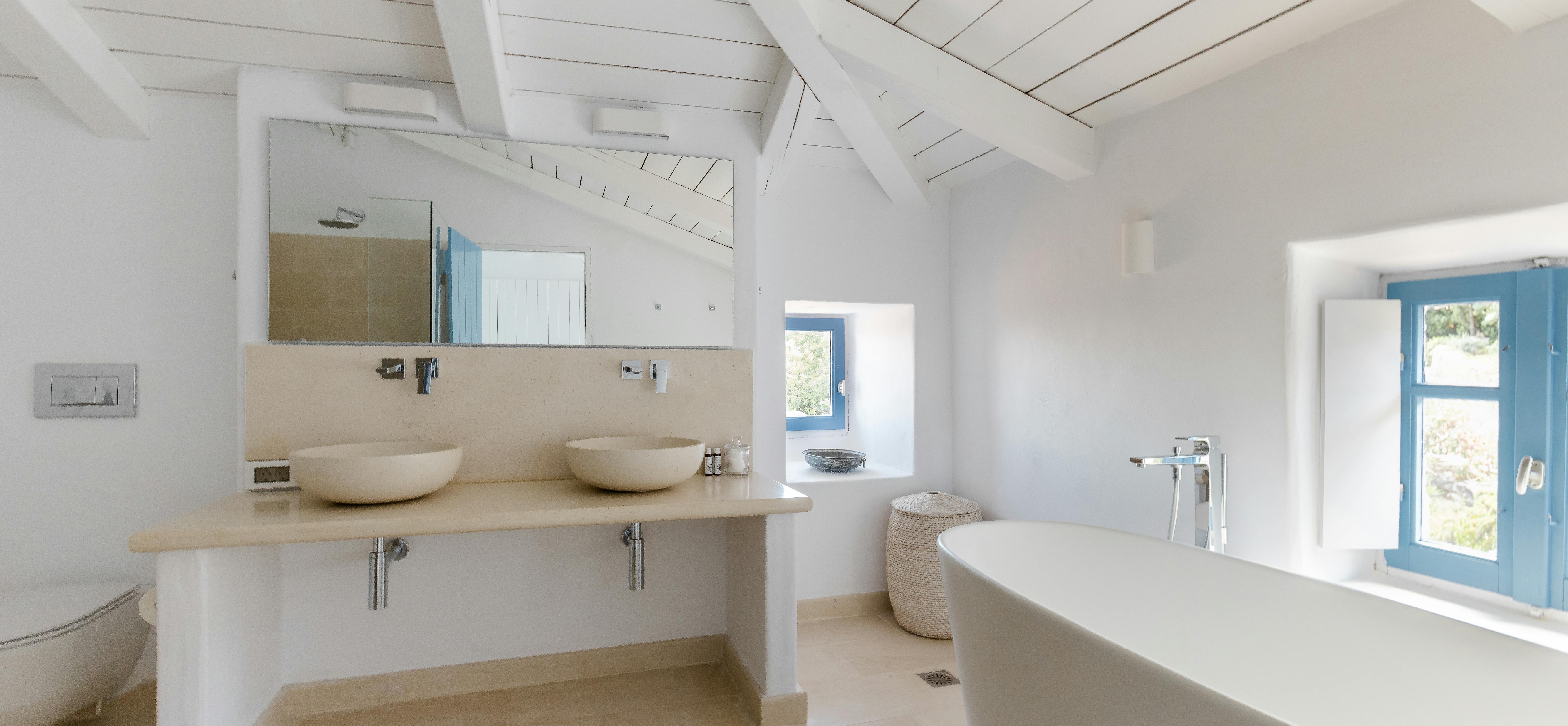 A serene white bathroom with vaulted wood ceiling, double vessel sinks on floating vanity, freestanding tub, and blue-trimmed windows.
