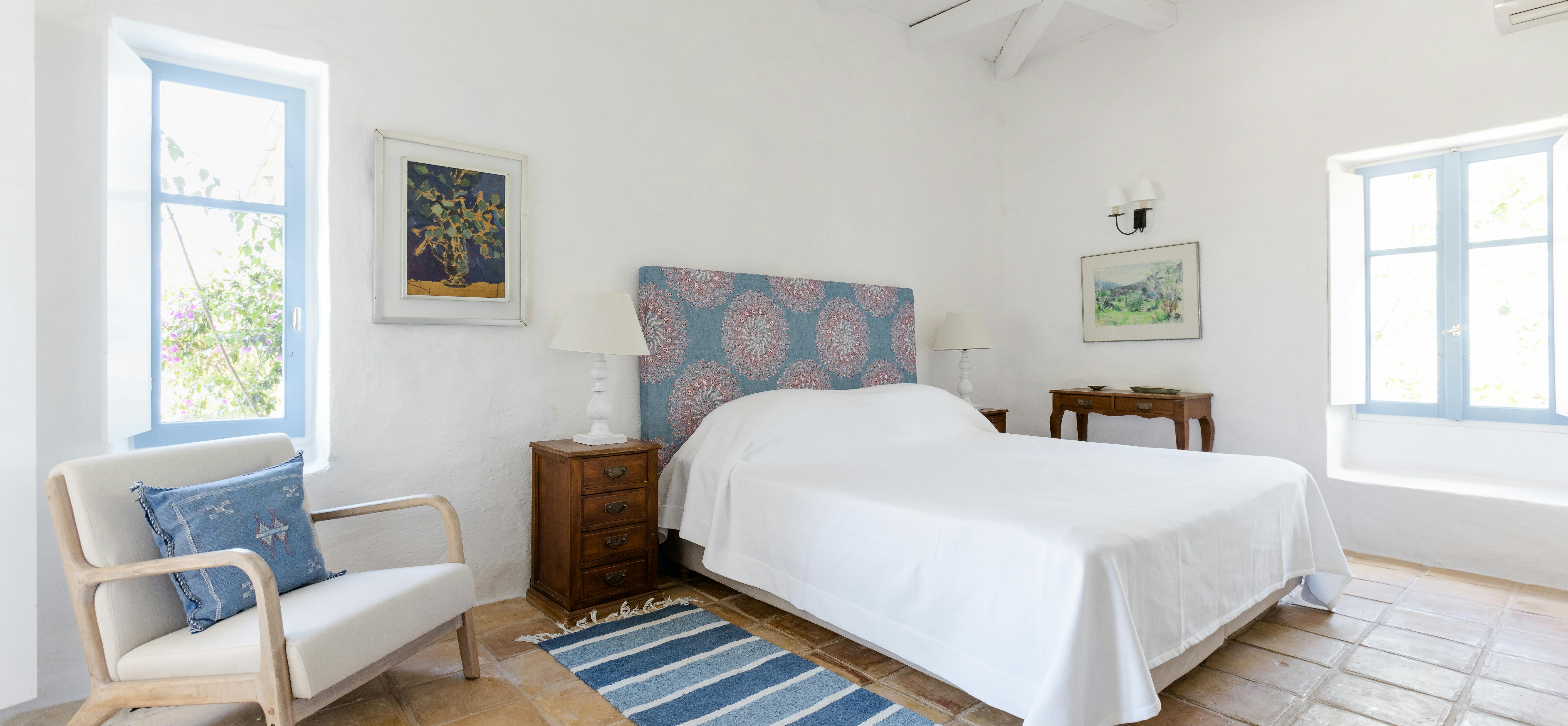A bright bedroom with white vaulted ceiling, blue and coral patterned upholstered headboard, white linens, cream armchair, and terracotta floors.