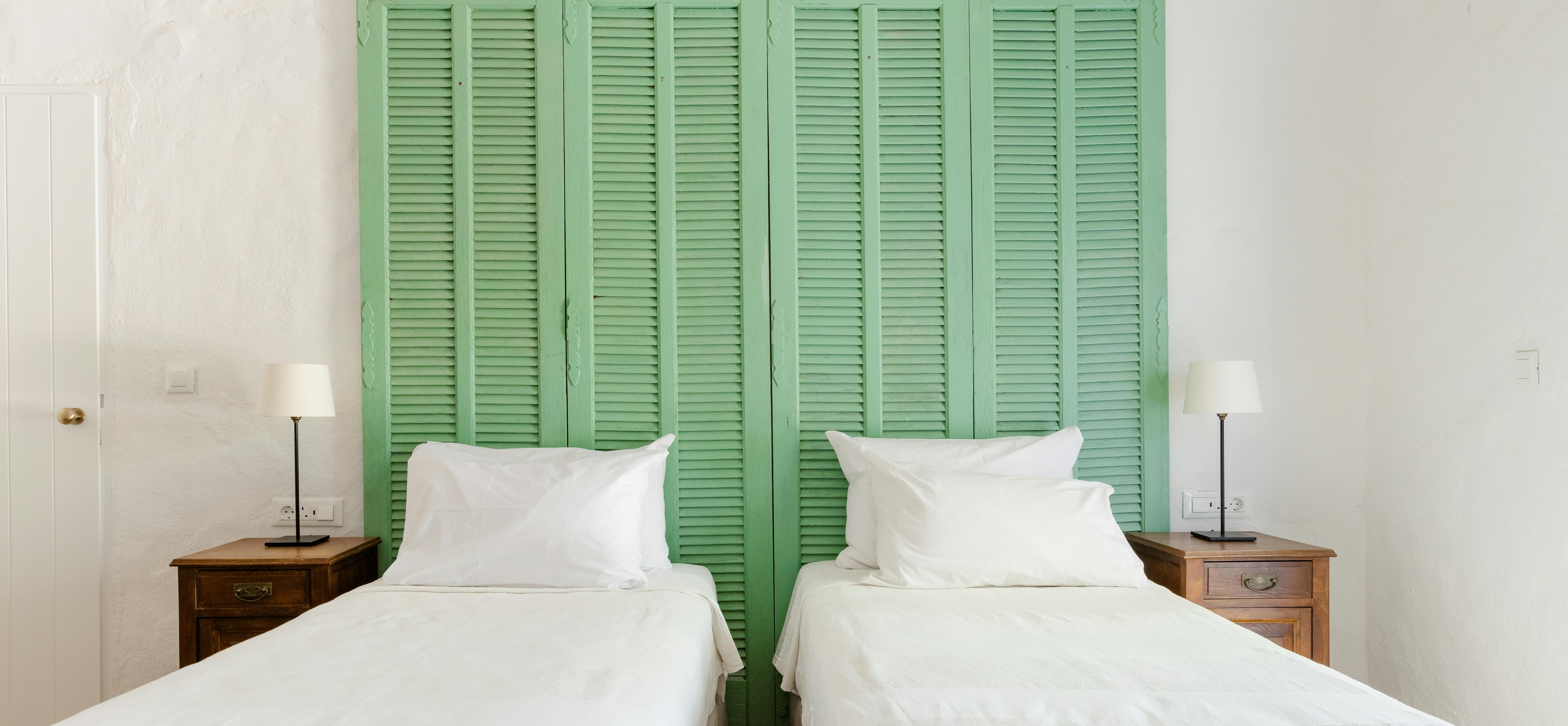 A symmetrical view of two twin beds with crisp white linens and mint green louvered shutters serving as headboards, flanked by matching wood nightstands.