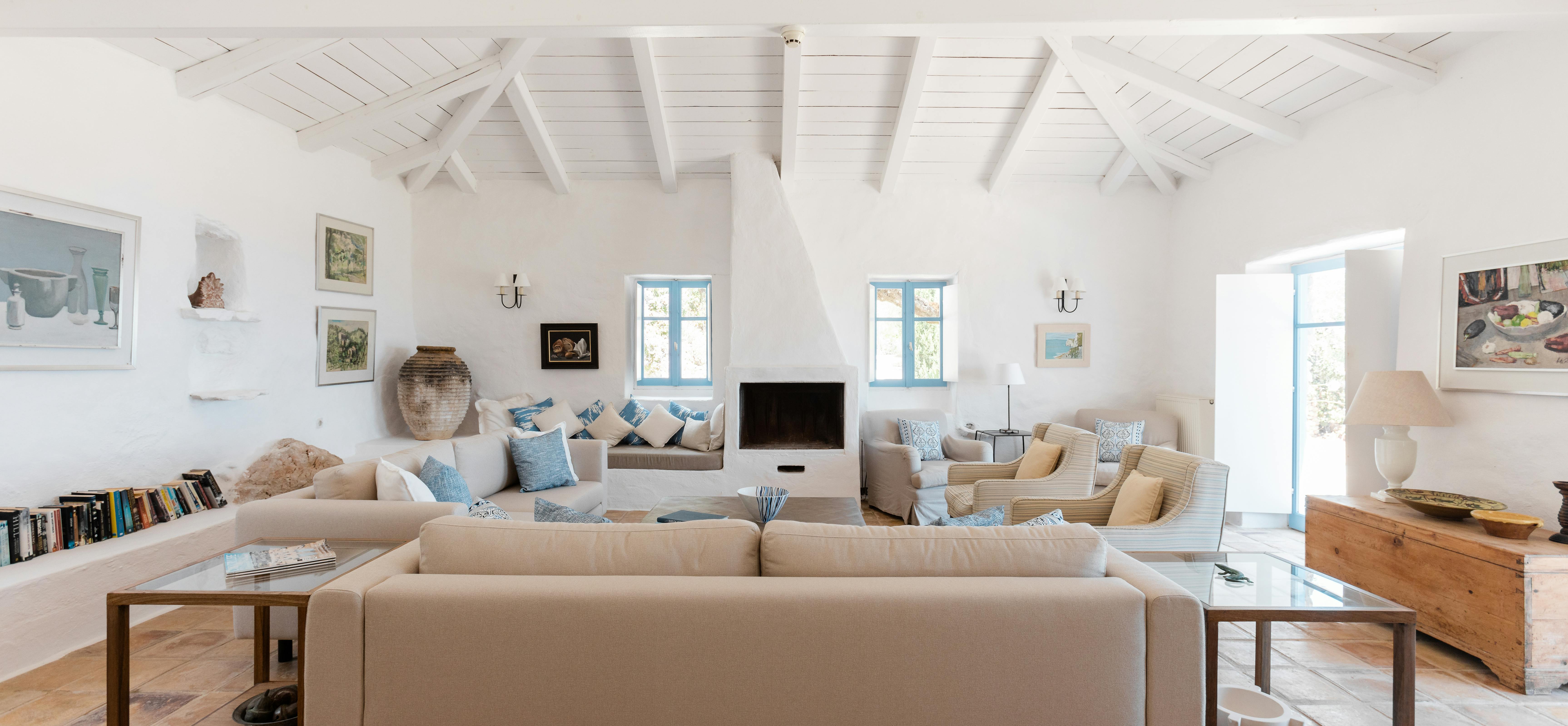 A spacious white living room with dramatic vaulted ceiling, multiple beige sectional sofas with blue accent pillows, modern coffee tables, and terracotta tile floor.