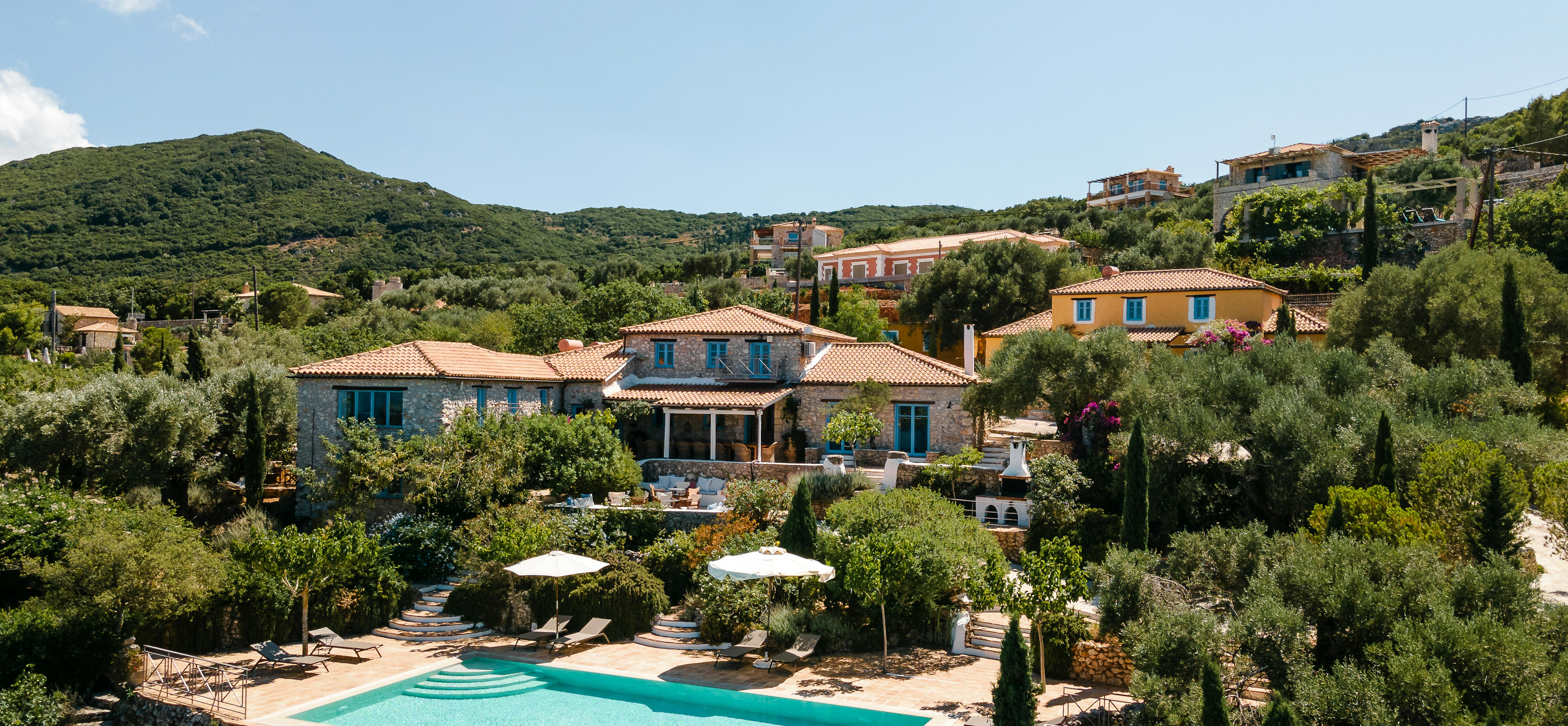 An aerial view of a Mediterranean stone villa complex with terracotta roofs, infinity pool with loungers, terraced gardens, olive trees, and hillside setting.