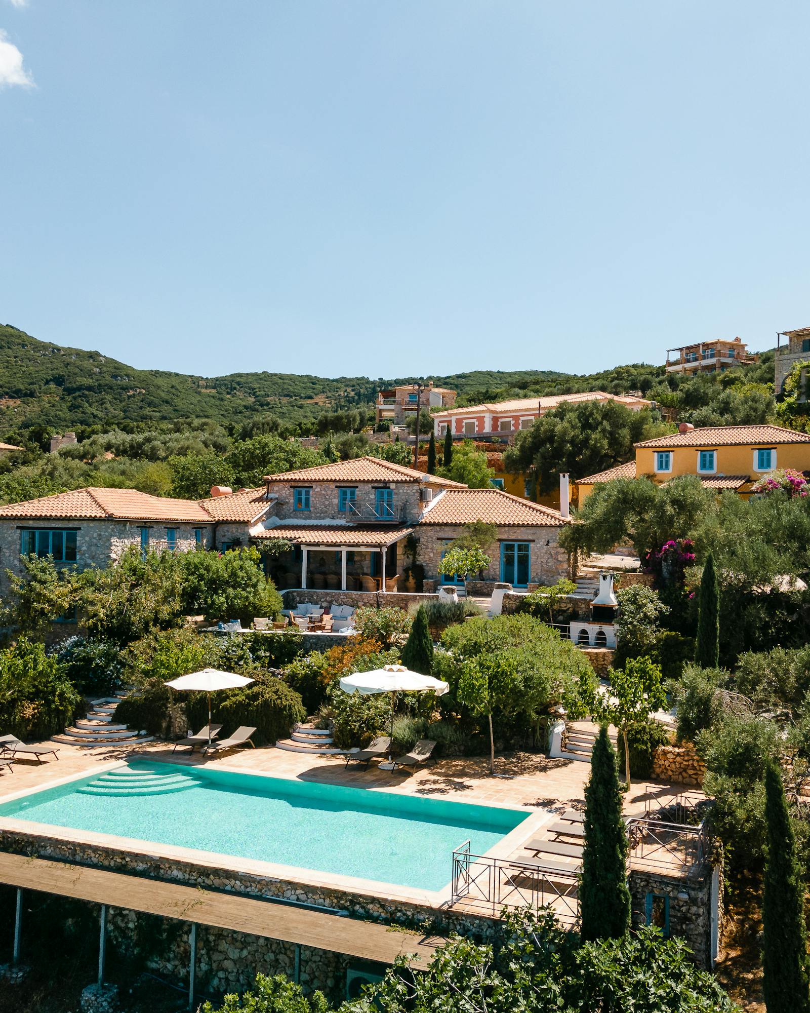 An aerial view of a Mediterranean stone villa complex with terracotta roofs, infinity pool with loungers, terraced gardens, olive trees, and hillside setting.