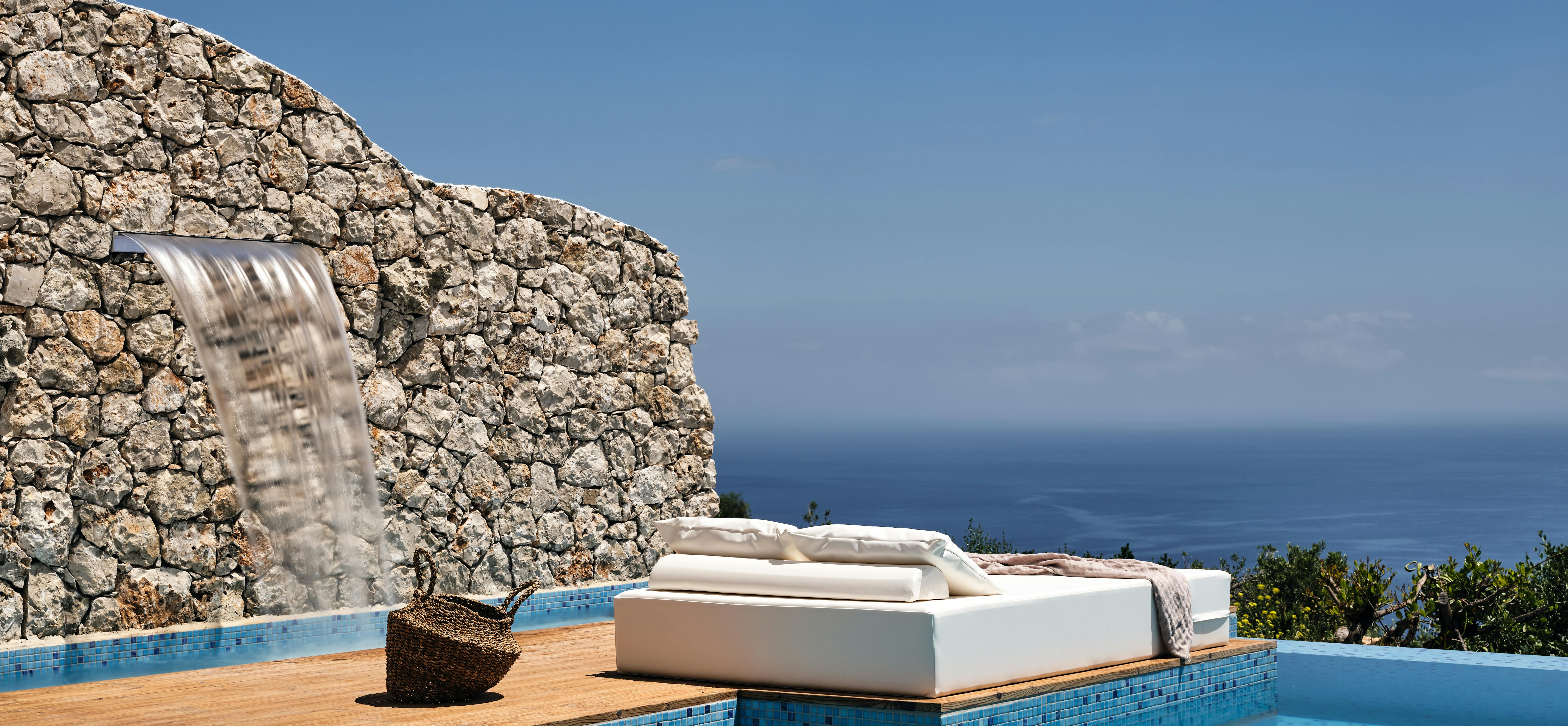 Infinity pool with integrated wooden platform supporting white loungers and waterfall feature against rugged stone wall with ocean beyond.