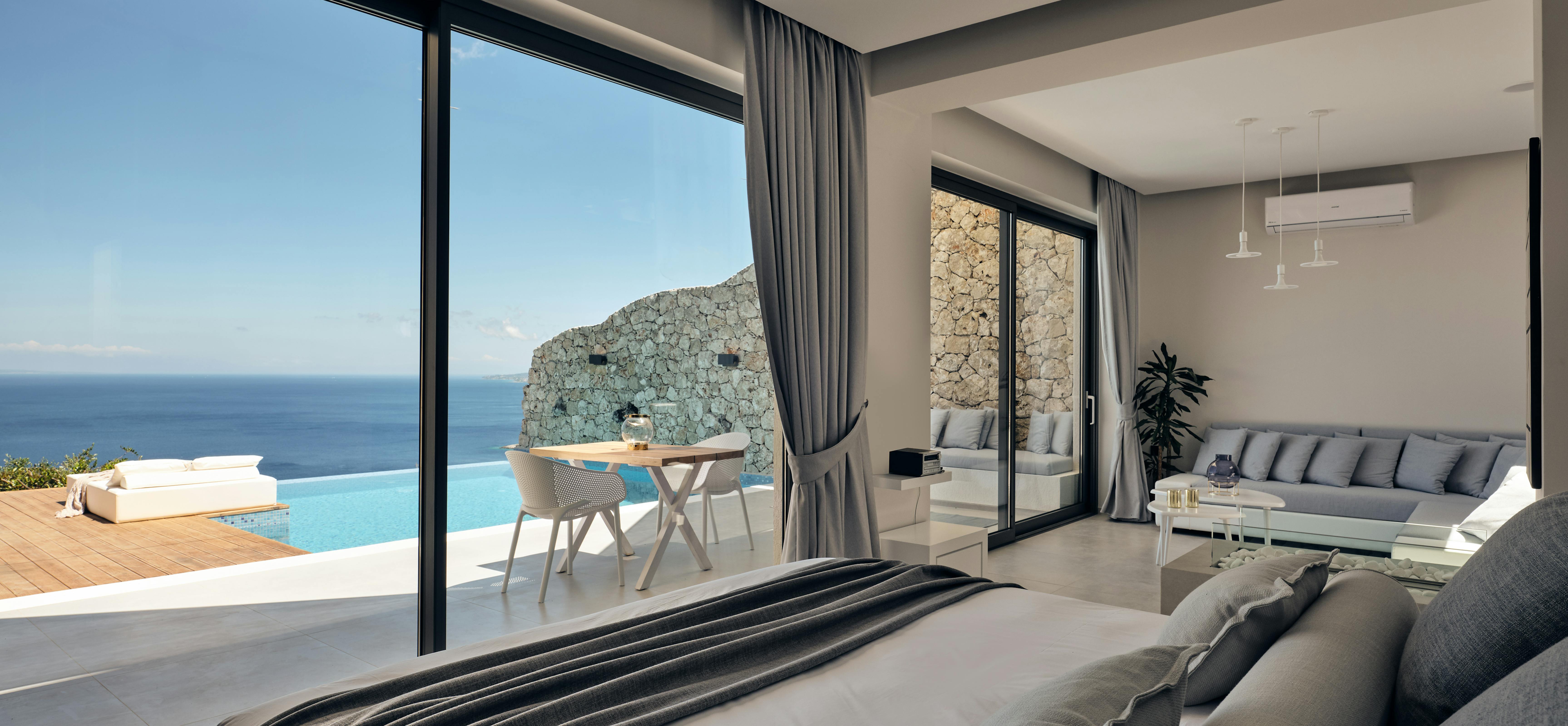 Spacious bedroom with ocean views through floor-to-ceiling windows, opening to a pool terrace with outdoor dining and stone wall backdrop.