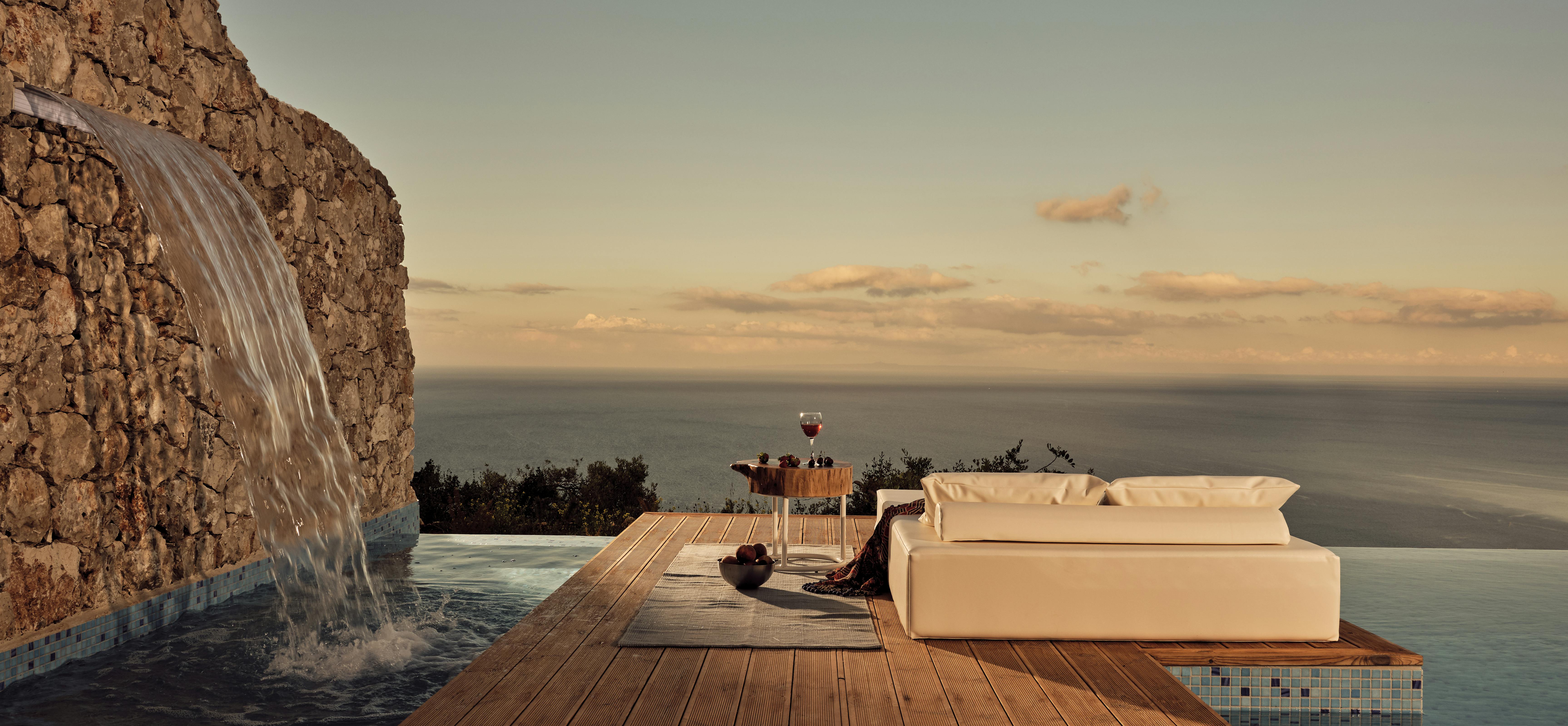 Sunset pool deck with stone waterfall feature, wooden platform with white cushioned daybed, and panoramic ocean views.