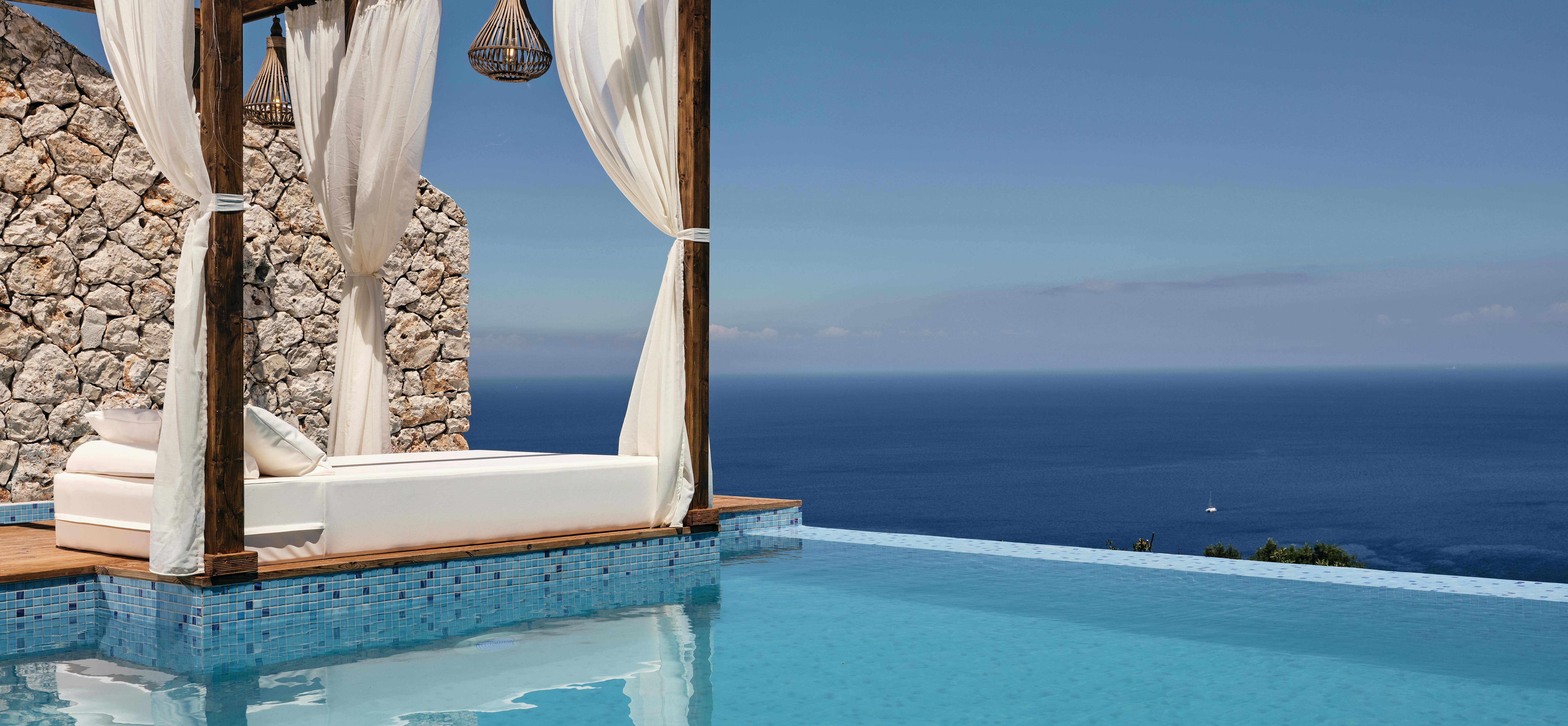Infinity pool with wooden cabana bed draped in white curtains and pendant lights, overlooking endless ocean horizon.