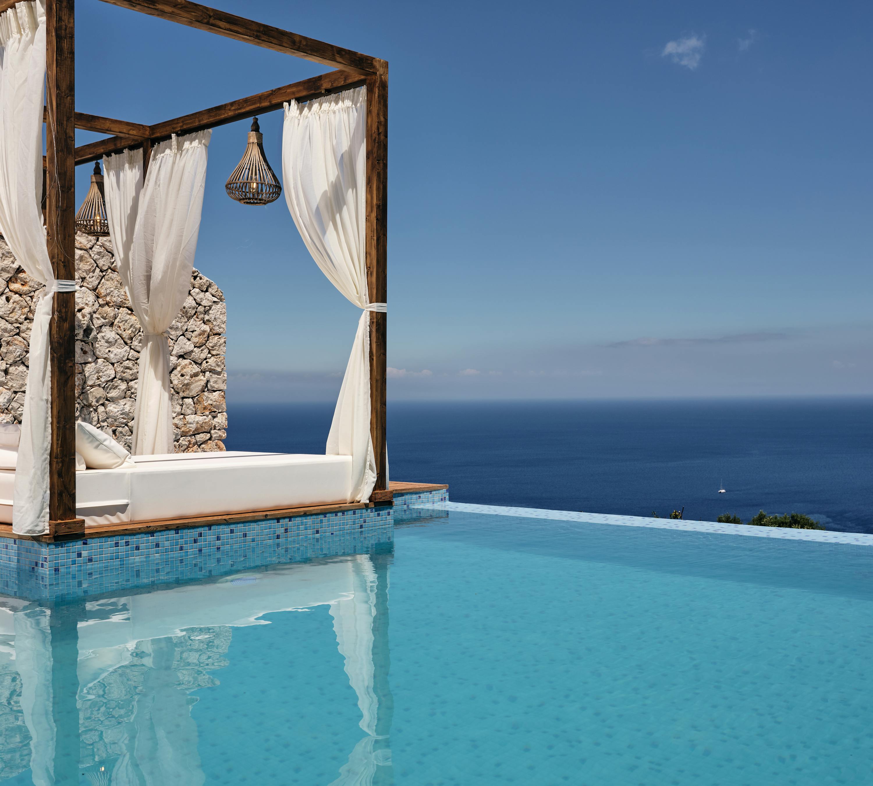 Infinity pool with wooden cabana bed draped in white curtains and pendant lights, overlooking endless ocean horizon.