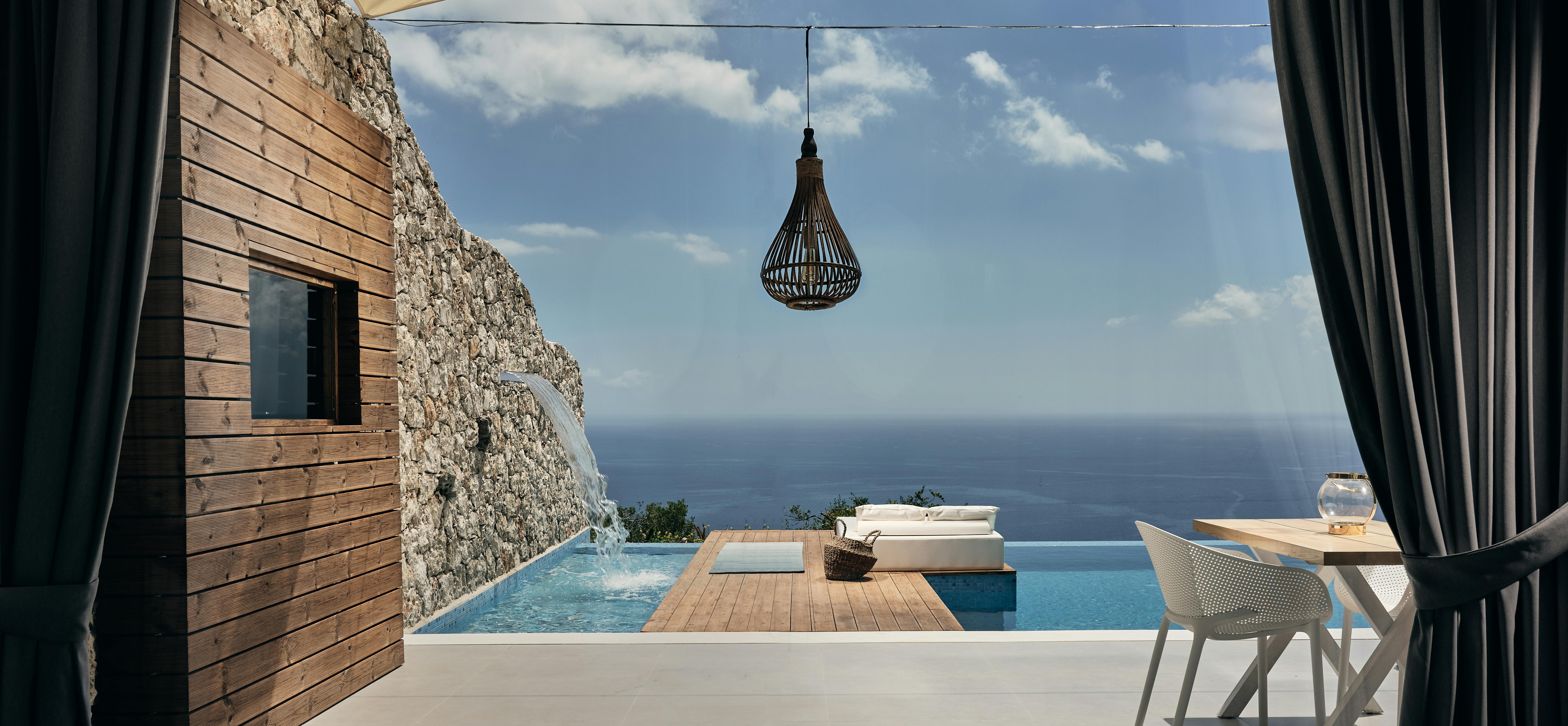 Bedroom view framing outdoor terrace with infinity pool, wooden deck, hanging lantern, and endless ocean horizon through dark curtains.