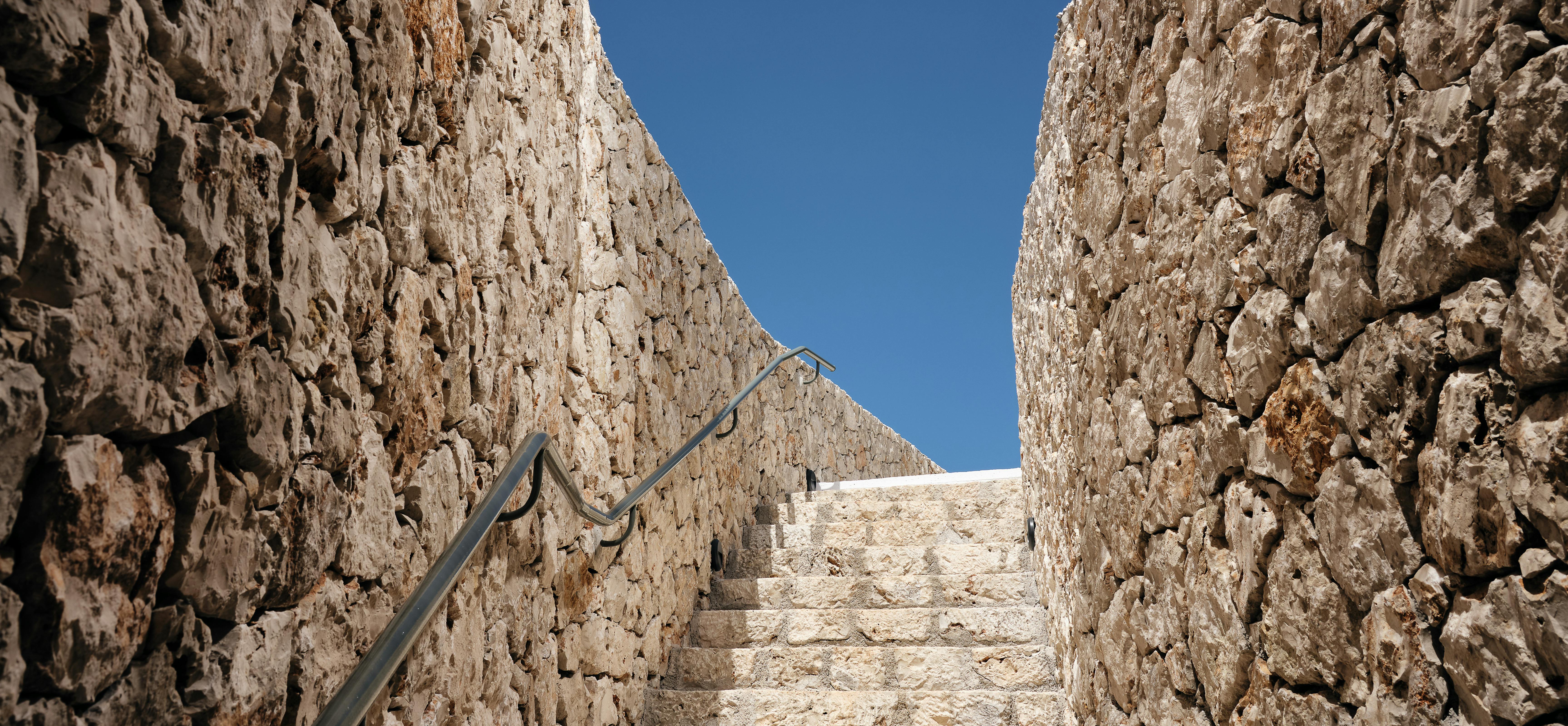 Stone stairway ascending between ancient limestone walls under a wooden pergola with clear blue sky visible above.